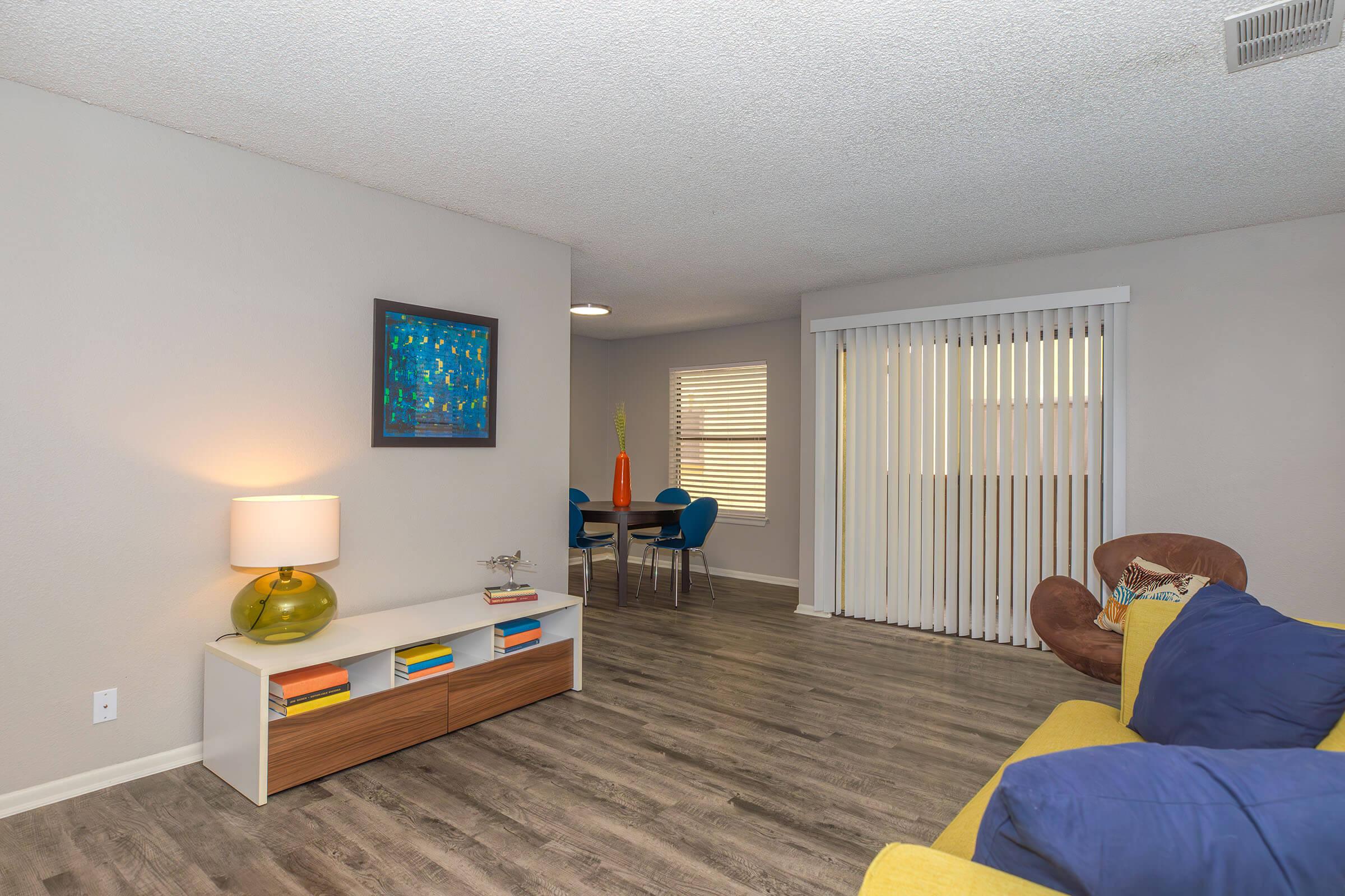 A spacious living room featuring a yellow couch with blue pillows, a modern coffee table, and a decorative lamp. A round dining table with blue chairs is visible in the background, along with a large window covered by vertical blinds. The walls are painted light gray, creating a bright and inviting atmosphere.