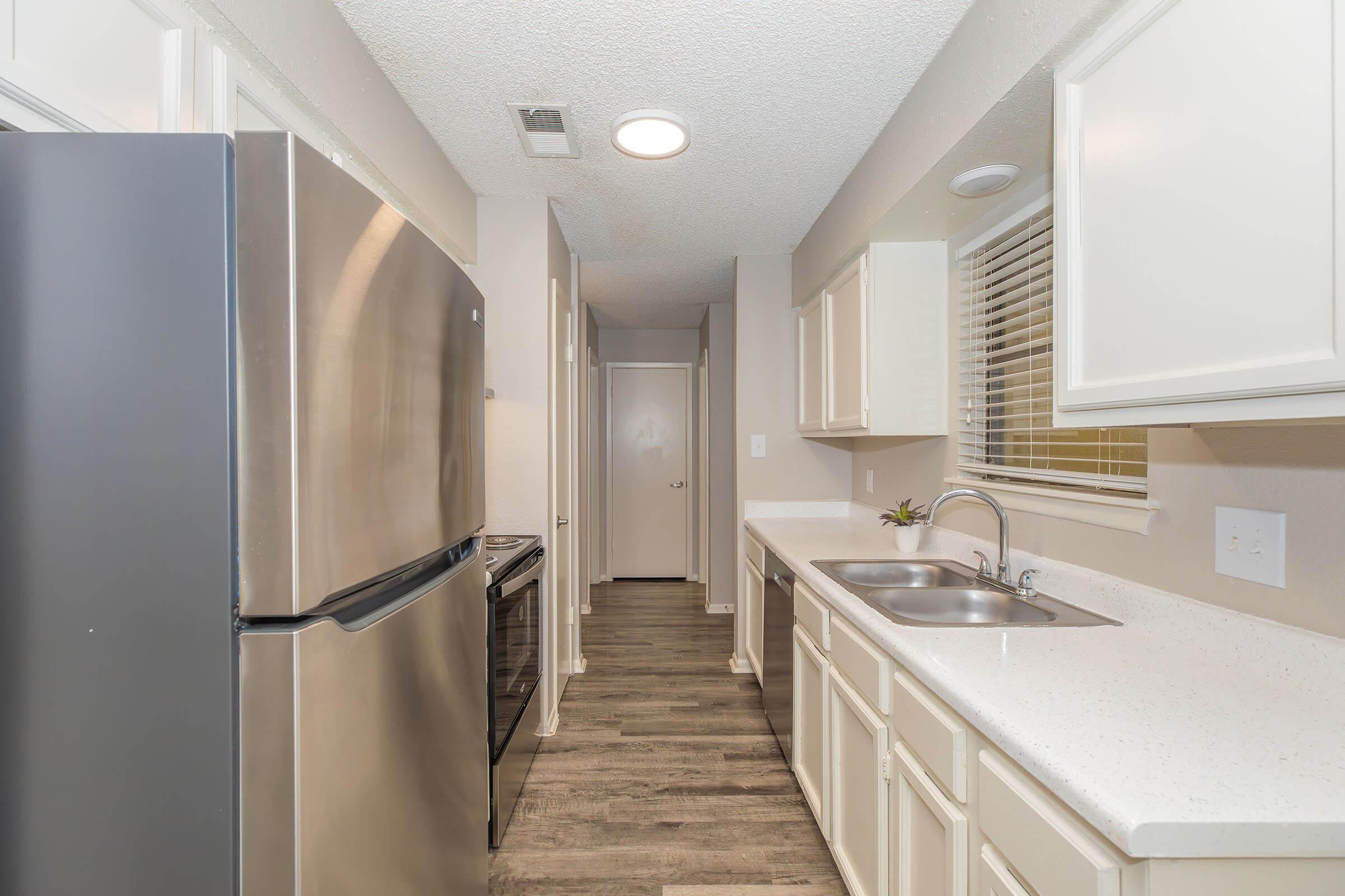 A modern kitchen with stainless steel appliances, including a refrigerator and stove. The countertops are white with a glossy finish, and the cabinets are light-colored. A small window allows natural light, and the flooring is a dark wood laminate. The kitchen appears tidy and well-lit.