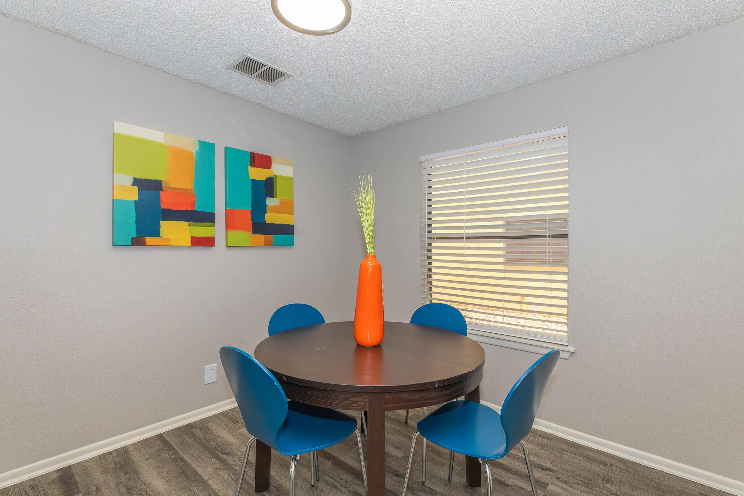 A cozy dining area featuring a round wooden table with four blue chairs. An orange vase with decorative plants is centered on the table. The walls are a light gray, and two colorful abstract paintings hang above the table, adding vibrancy to the space. A window with blinds lets in natural light.