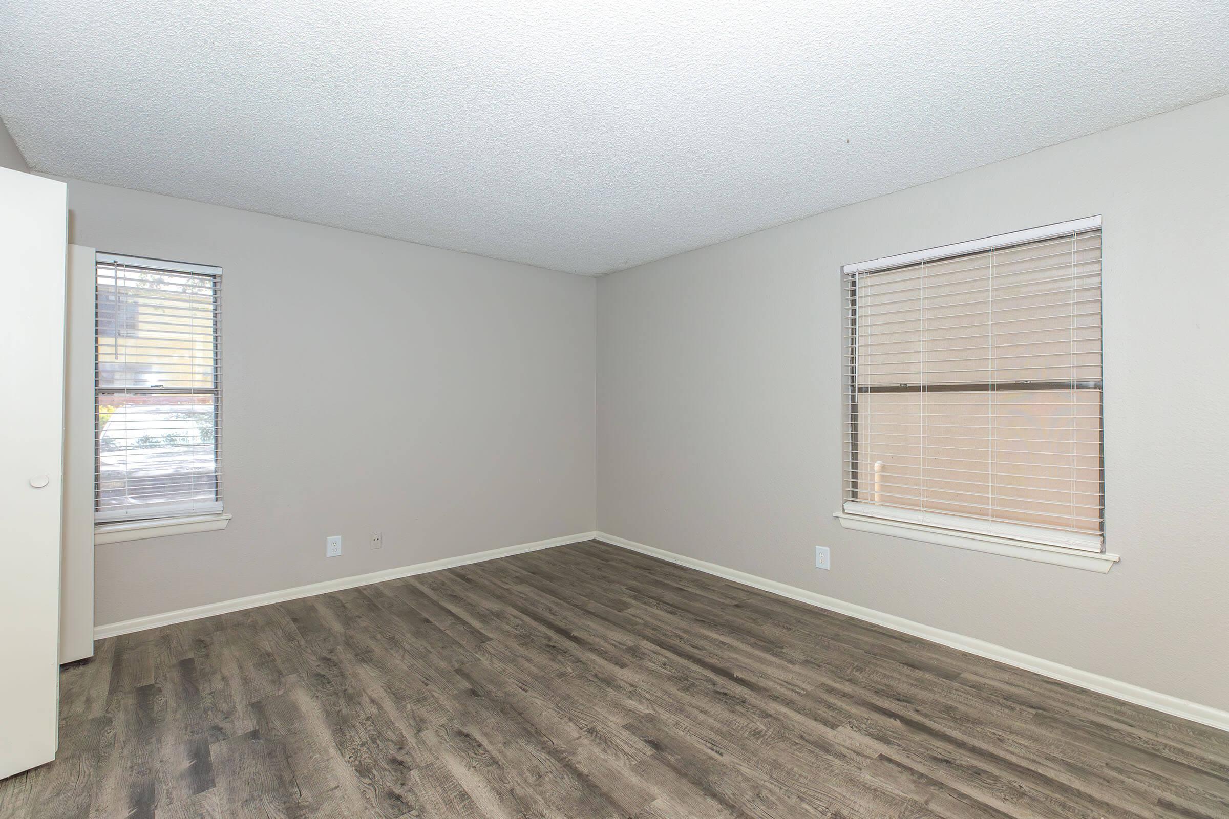 Empty room with light gray walls and hardwood flooring. Two windows with blinds allow natural light, and a closet door is visible on one side of the room. The overall atmosphere is neutral and spacious, suitable for various decorative styles.