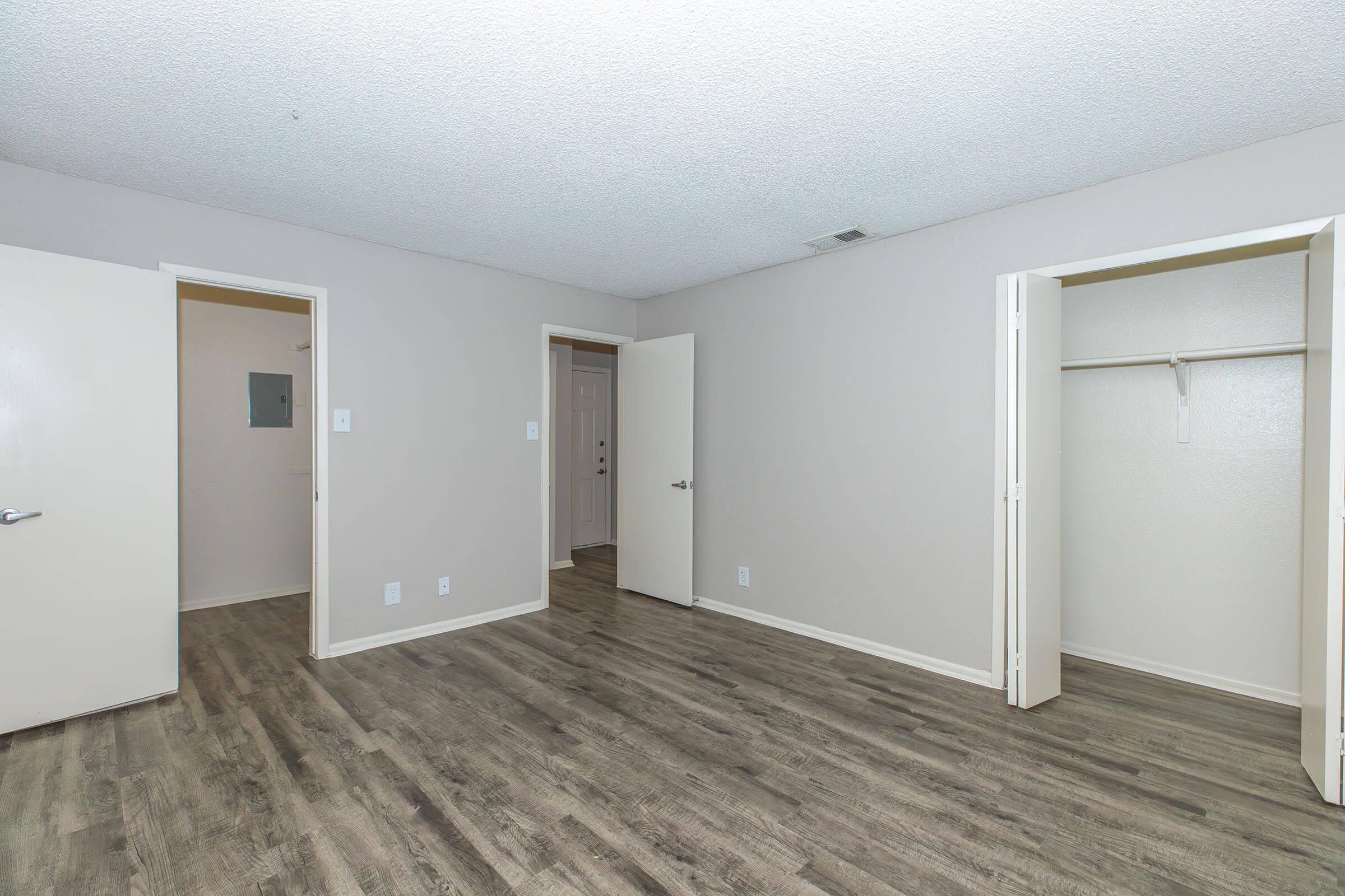 Empty room with light gray walls and new laminate flooring. Two white doors and an open closet with a hanging rod are visible. The room has a bright and spacious feel, with a ceiling that appears textured. Natural light illuminates the space.