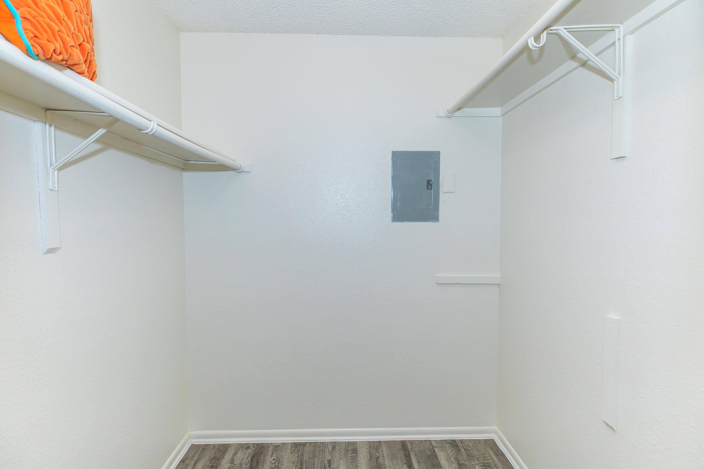 A vacant closet with white walls and two hanging shelves. The flooring is wood-like laminate. There is a small electrical panel on one wall, and an orange storage item is placed on the top shelf. The space is well-lit and uncluttered, suggesting it is ready for organization or storage.