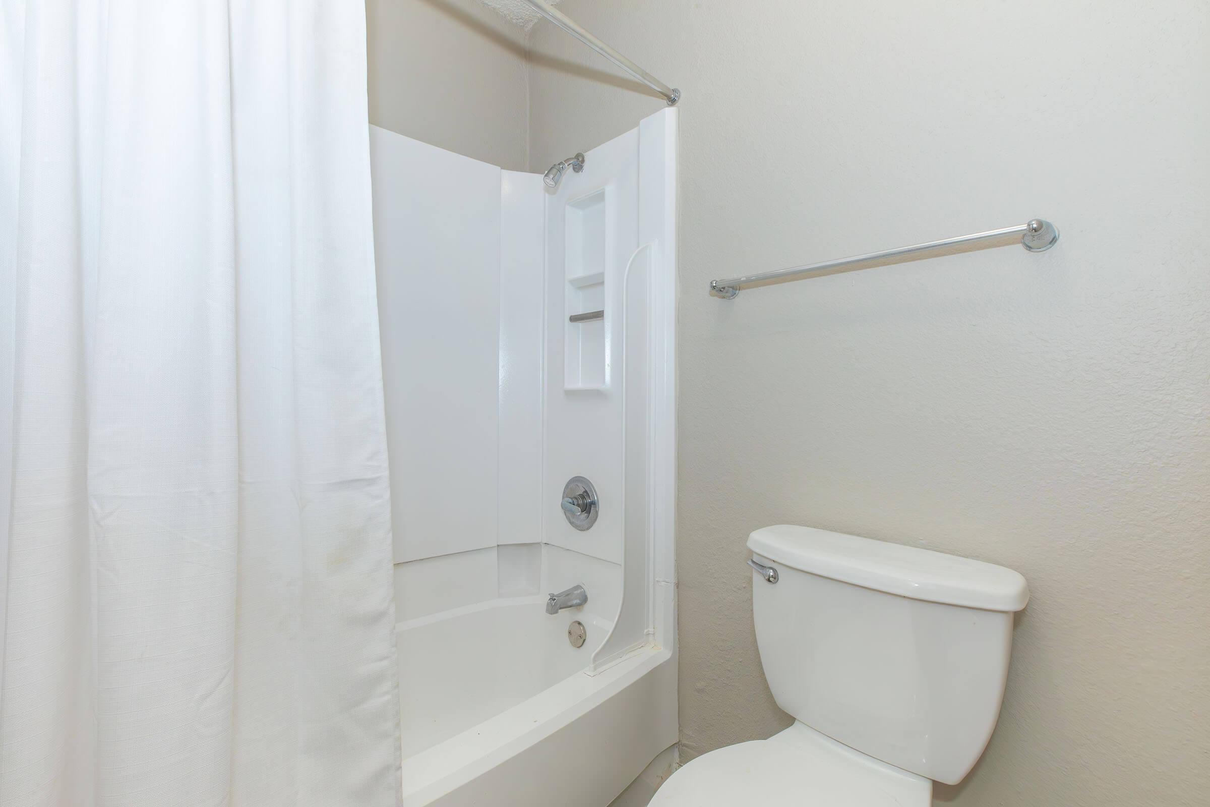 A clean bathroom with a white bathtub and shower combination, featuring a shower curtain. To the right, there is a white toilet. The walls are light-colored, creating a bright and spacious appearance.