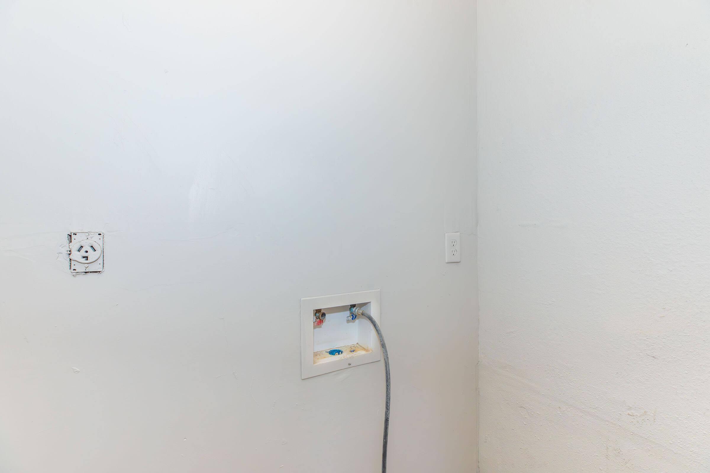 A plain white wall in a room with two water supply connections visible, one on the left and another on the right. The area appears unfinished, with a minor electrical outlet on the wall. The background is empty and uncluttered, creating a simple, minimalistic appearance.