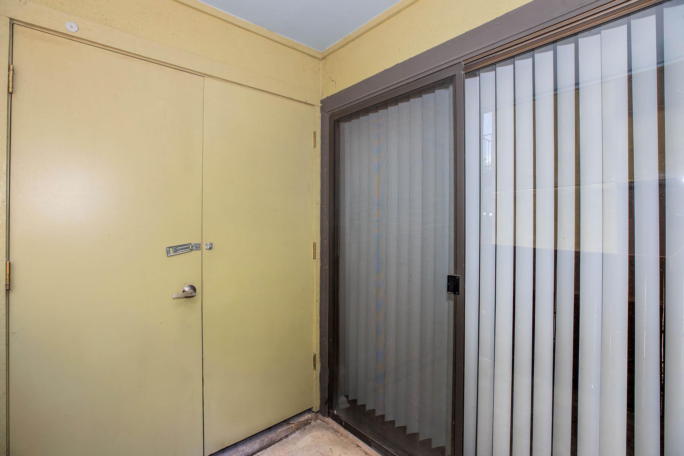 A small entryway with a beige wall and floor. On the left, there is a closed door with a small window and a handle. To the right, a sliding glass door with vertical blinds partially open, leading to an outdoor area. The space appears bright and well-maintained, with neutral colors and minimal decor.
