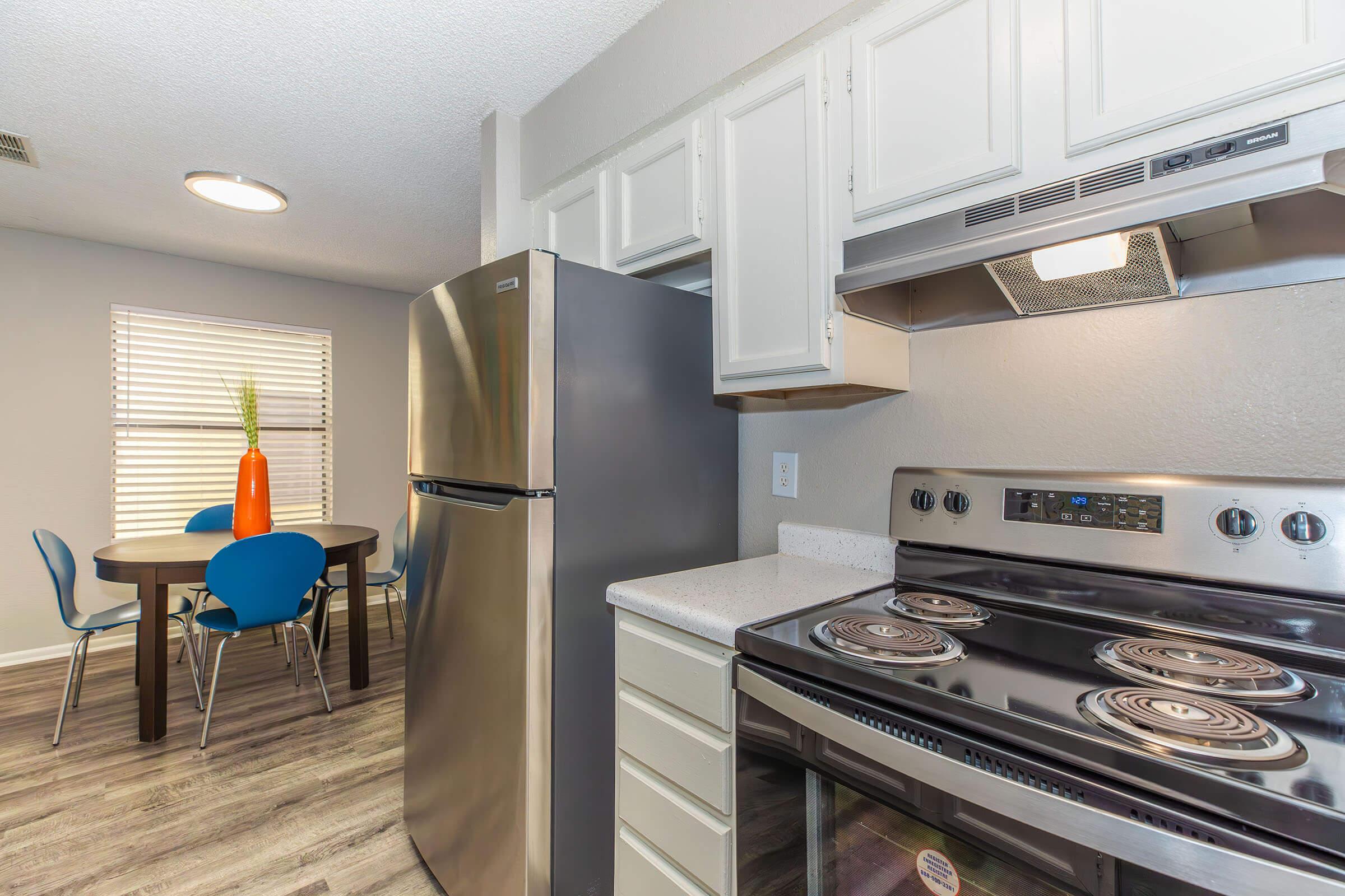 Modern kitchen featuring stainless steel appliances, including a fridge and stove. A round dining table with four blue chairs sits near a window with blinds. Light wood flooring adds warmth to the space, complemented by neutral wall colors and a decorative plant in a vase on the table.