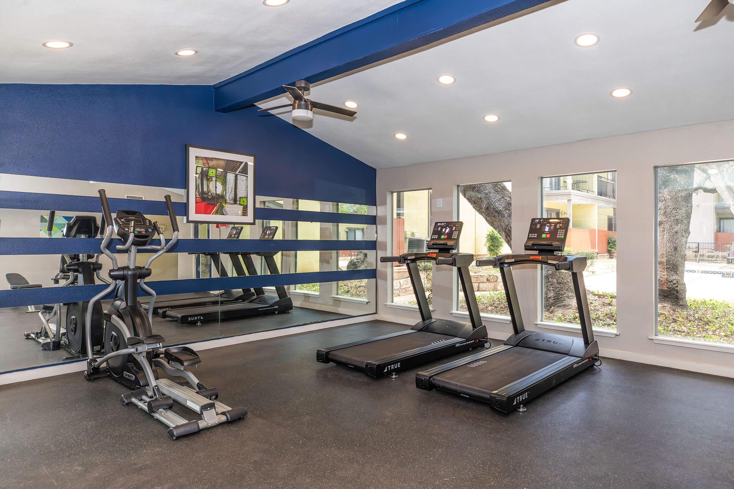 Interior view of a modern gym featuring two treadmills and an elliptical machine. The walls are painted blue and there are large windows that allow natural light to fill the space. A mirror stretches along one wall, reflecting the equipment and the outdoor scenery.