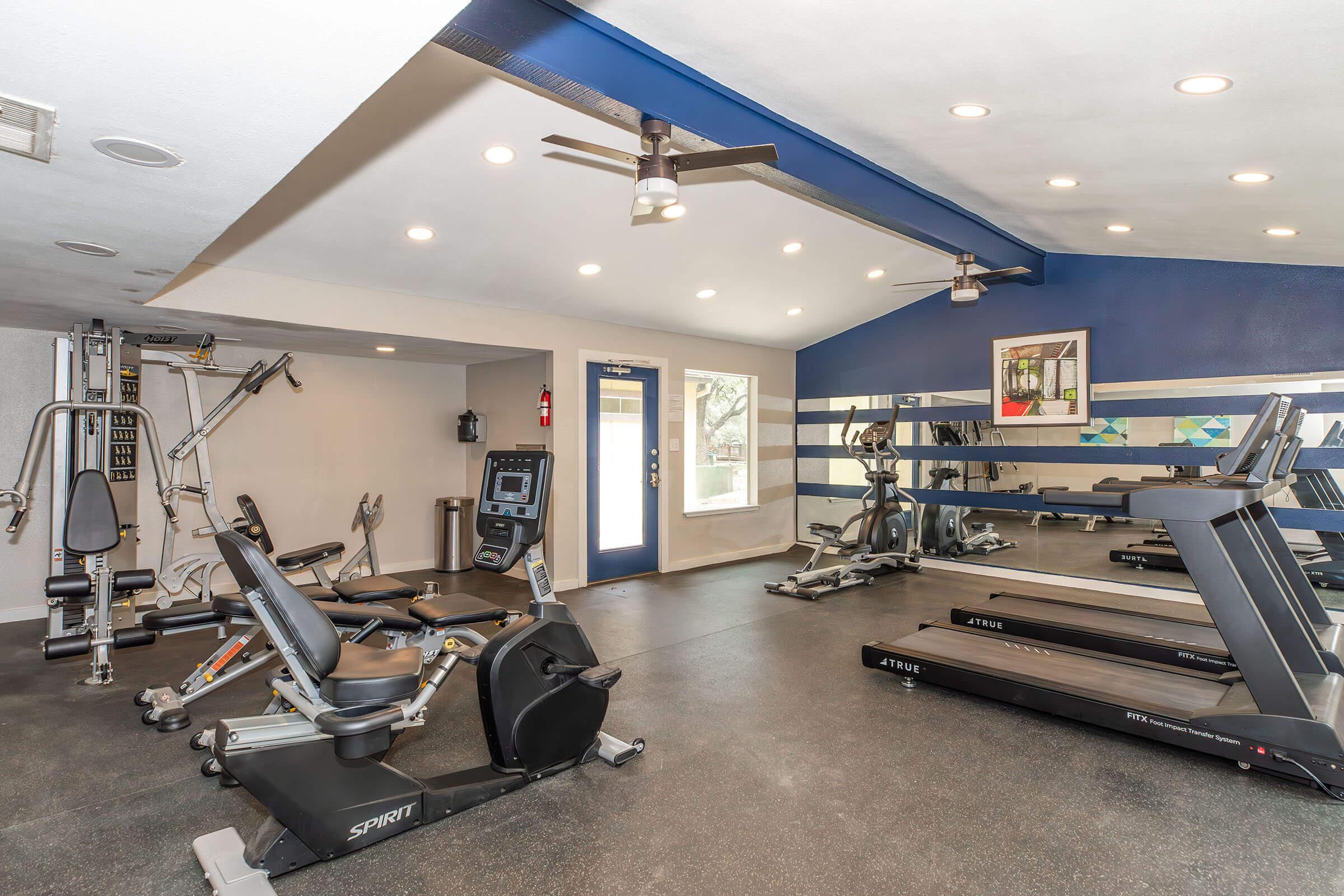 A well-lit gym area featuring various exercise equipment including treadmills, a weight machine, and a stationary bike. The walls are painted in blue and white, with a large window and modern ceiling fans providing ventilation. The space has a clean and organized appearance, ideal for workouts.