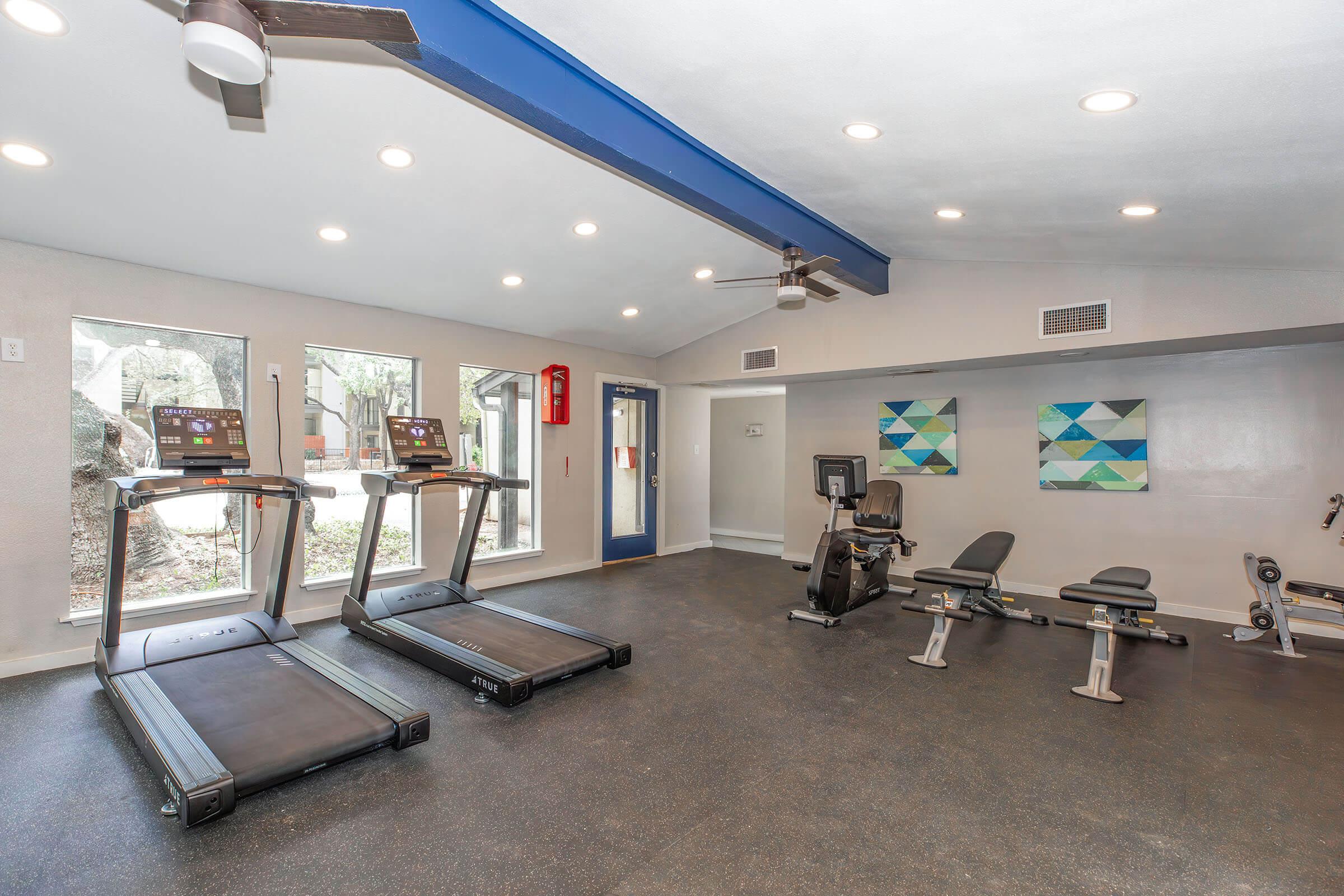 A modern gym interior featuring two treadmills, an exercise bike, and a weight bench. Large windows allow natural light to brighten the space, which is decorated with abstract geometric art on the walls. The floor is covered with dark rubber flooring for safety and comfort.