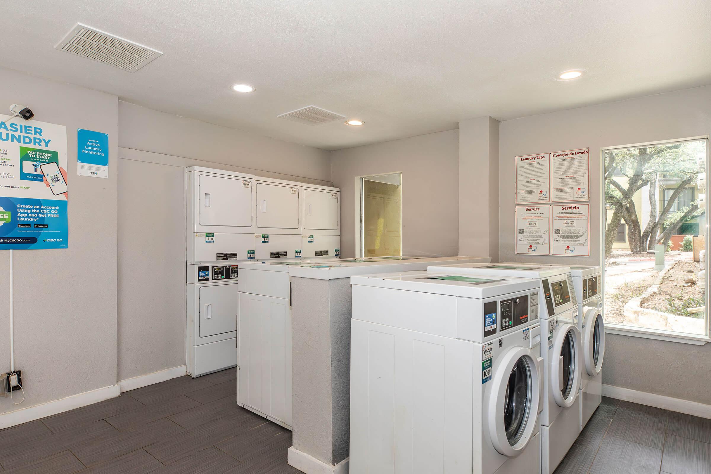 A bright and spacious laundry room featuring multiple washers and dryers. The machines are lined up against a light gray wall, with informational posters nearby. Large windows allow natural light to filter in, and there are trees visible outside, adding to the welcoming atmosphere.