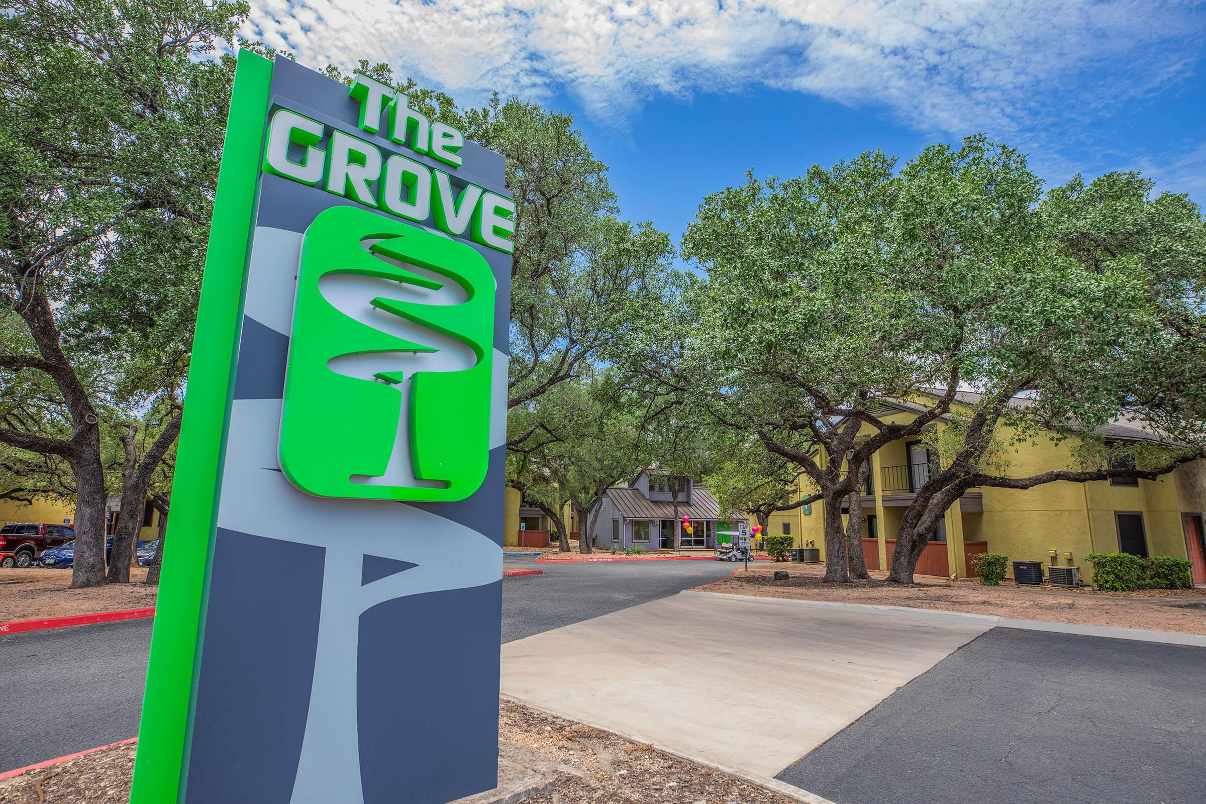 Sign for "The Grove" featuring a modern design, surrounded by trees and a clear blue sky. The sign is prominently displayed at the entrance of a multi-building complex that appears to offer accommodation, with parking spaces and pathways visible in the foreground.
