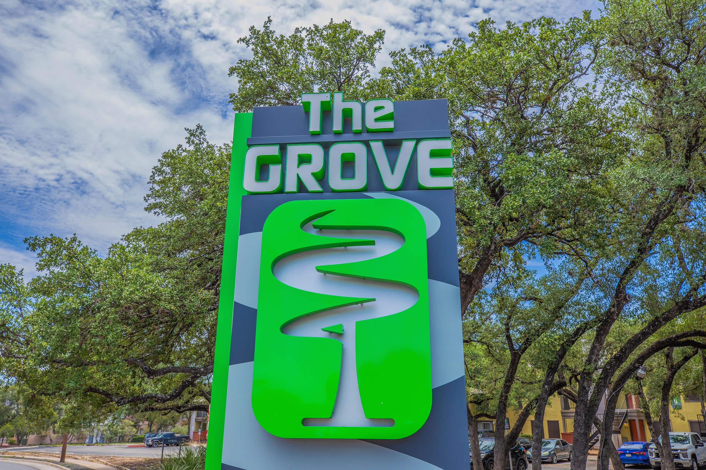 Sign for "The Grove" featuring a stylized green logo with a swirl design and a tree motif. The background includes a cloudy blue sky and surrounding greenery, suggesting a tranquil outdoor setting. The sign is prominently displayed in a landscaped area.