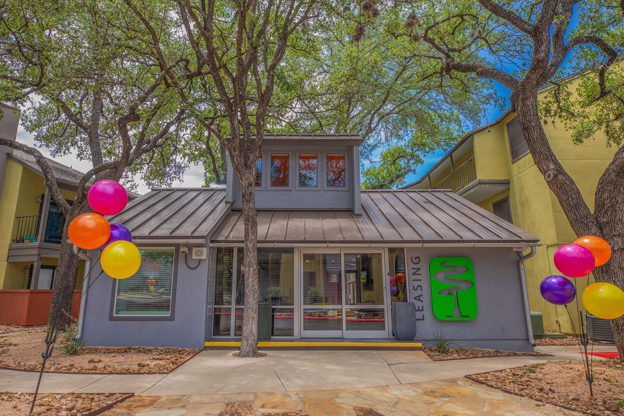 A leasing office with a modern facade surrounded by trees. The entrance features large windows and a green sign that says "LEASING." Colorful balloons in purple, orange, and yellow are tied to pillars on either side of the walkway, adding a festive touch to the outdoor area.