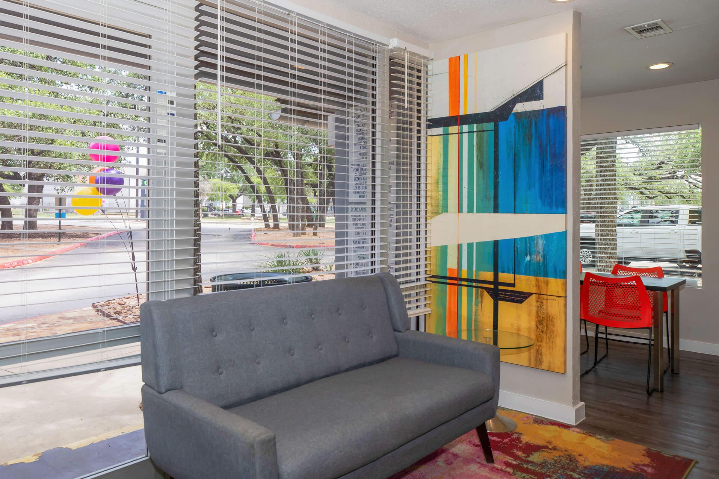 A modern interior space featuring a gray sofa, colorful abstract wall art, and large windows with blinds. Natural light fills the room, showcasing a dining area with orange chairs. Outside, greenery and colorful balloons are visible through the windows, adding a cheerful atmosphere.
