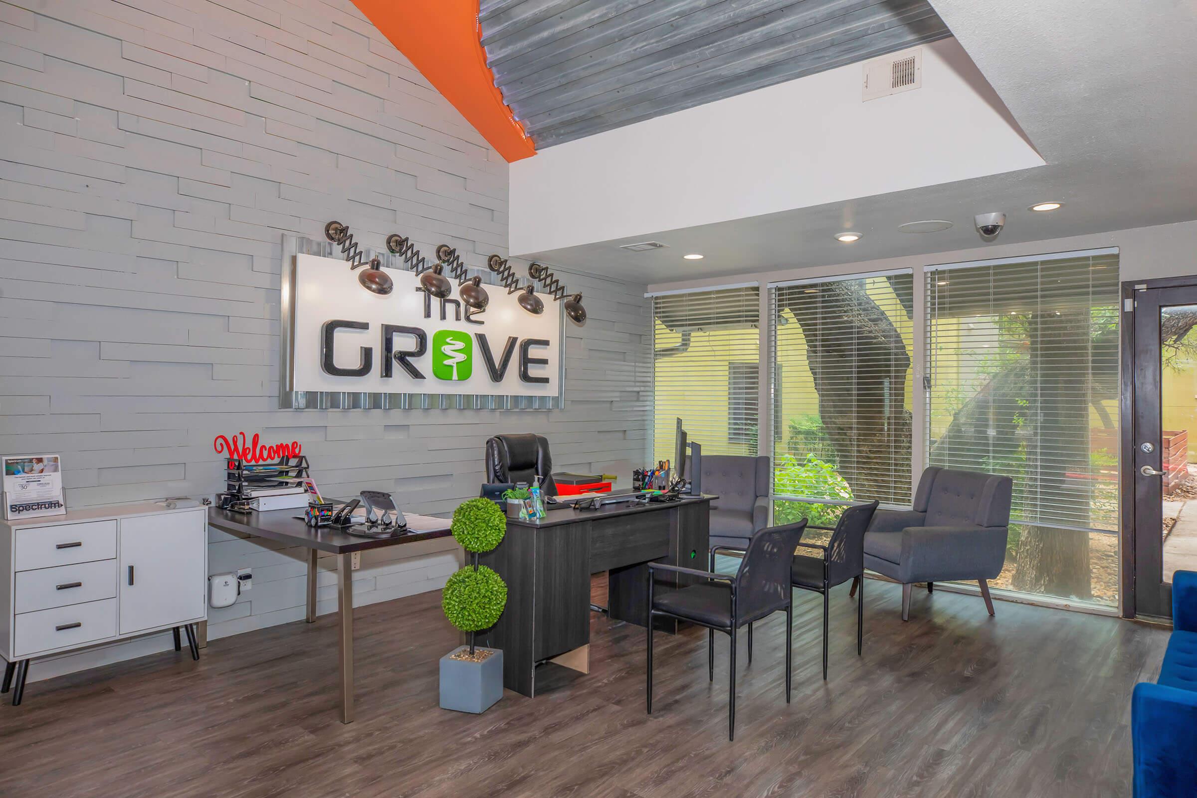 A modern office interior featuring a reception desk with the name "The Grove" displayed prominently. The space includes a stylish chair, a welcome sign, potted greenery, and large windows showing a view of trees outside. The decor features a mix of wood and gray walls with vibrant orange accents.