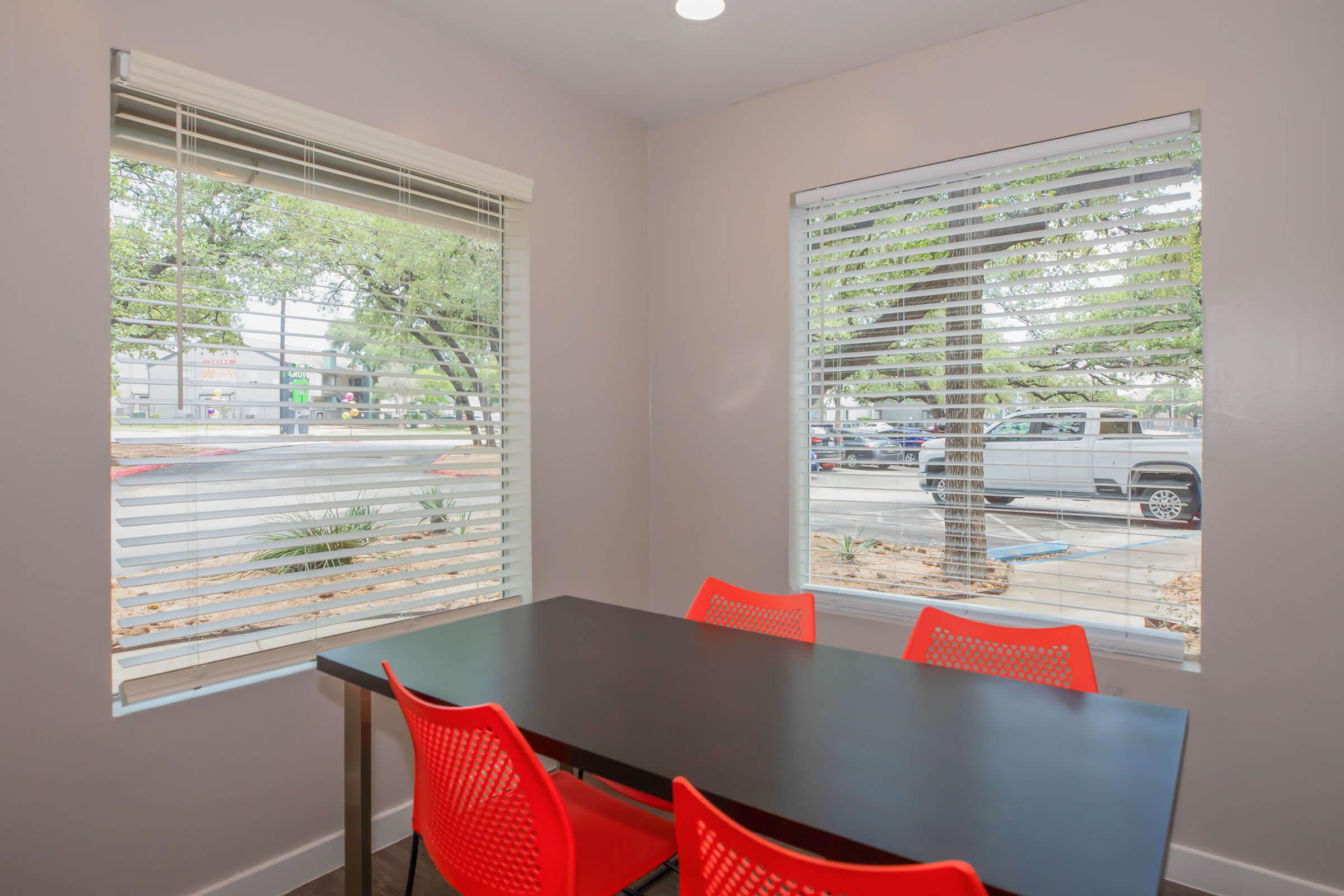 A small meeting room featuring a black table surrounded by four bright red chairs. There are two windows with blinds that allow natural light to enter, showcasing a view of trees and parked cars outside. The room has a modern, minimalist design with neutral-colored walls.