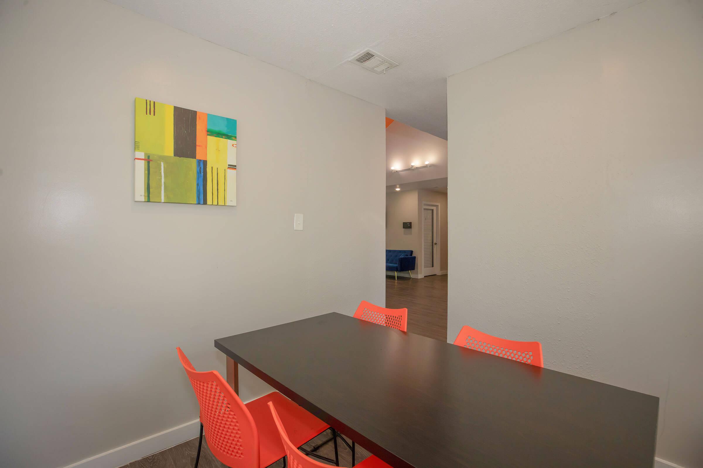 A dining area featuring a dark wooden table and four bright orange chairs. A colorful abstract painting hangs on the wall above the table. The space has light-colored walls and a view of a living room area in the background, which includes blue seating.