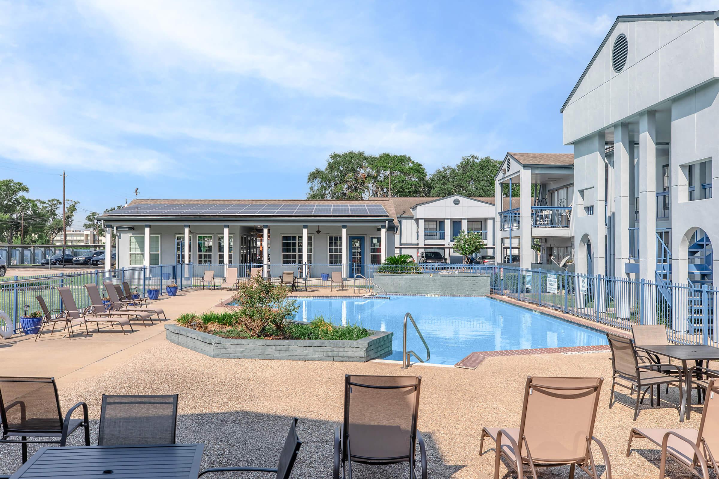 A swimming pool surrounded by lounge chairs, with a contemporary building in the background. The area features landscaping with plants and a blue sky overhead. The pool is inviting, with a clear blue water surface, and there are tables and chairs for relaxation.