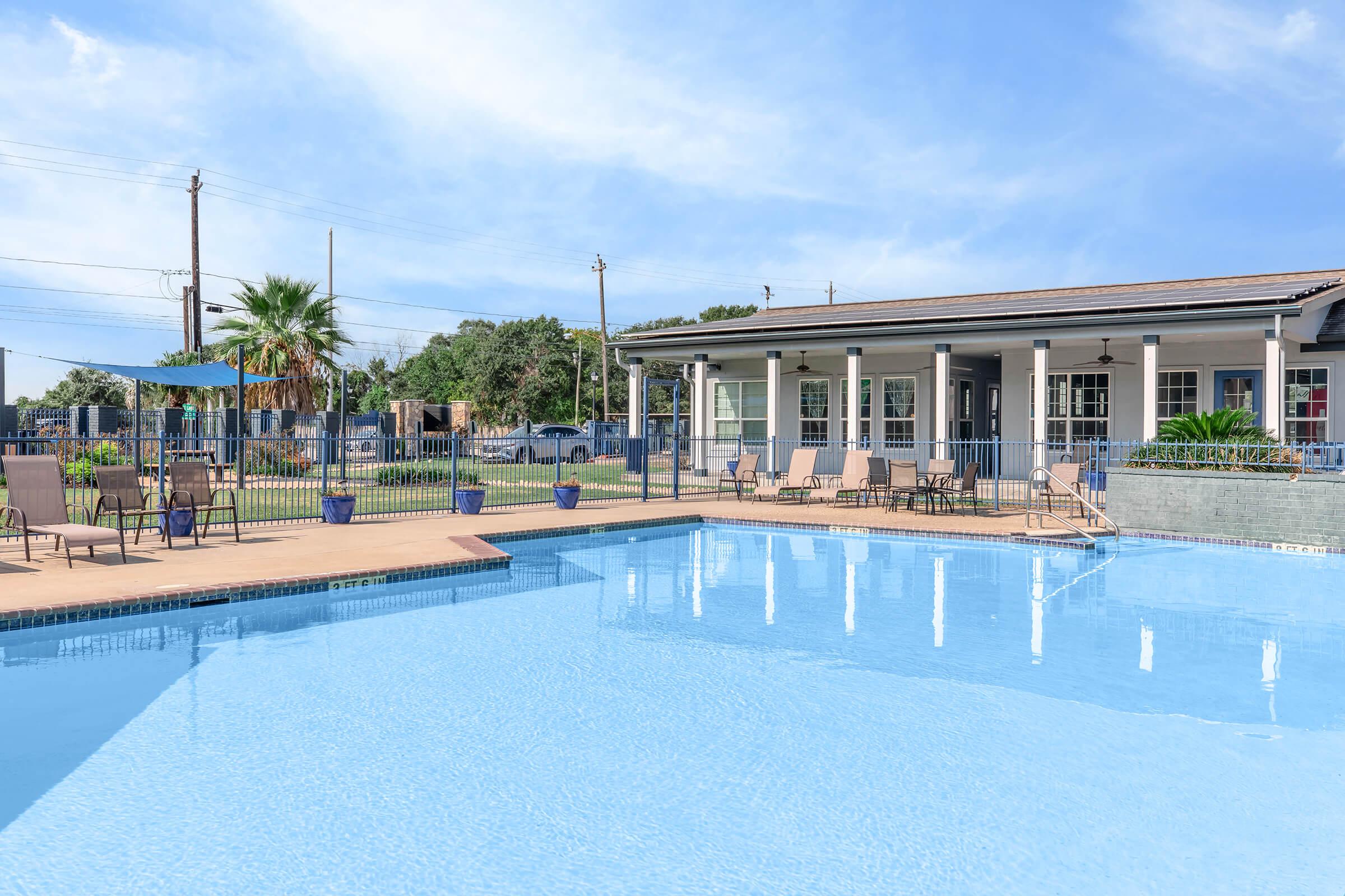A clear pool surrounded by lounge chairs and umbrellas, with a modern building in the background. The scene is set under a bright blue sky, showcasing a relaxing outdoor space perfect for leisure activities. Green trees and a landscaped area enhance the ambiance.