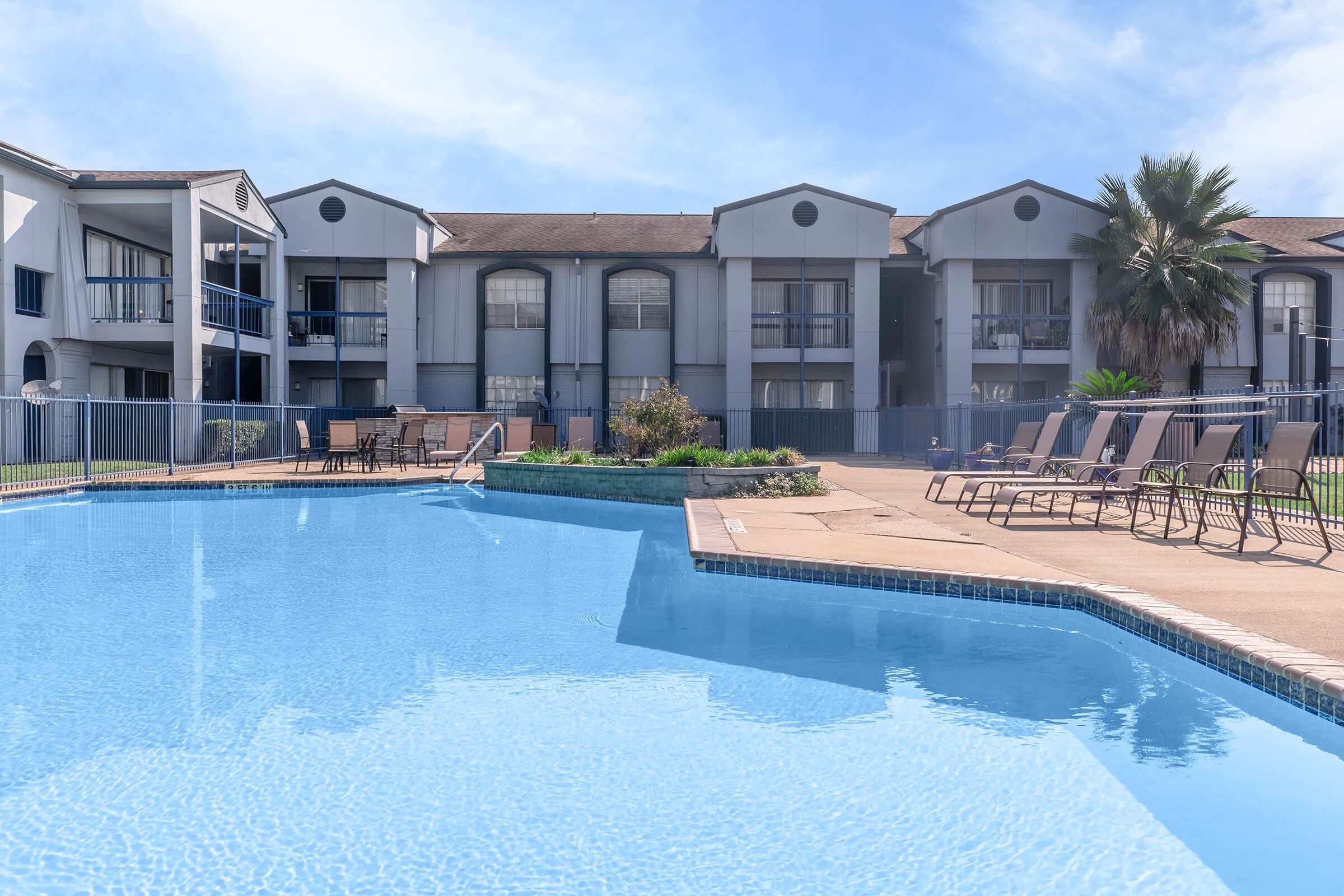A clear blue swimming pool in the foreground, with lounge chairs arranged around it. In the background, a multi-story apartment complex is visible. The sky is bright and sunny, with a few clouds. A small garden area with greenery is situated near the pool.