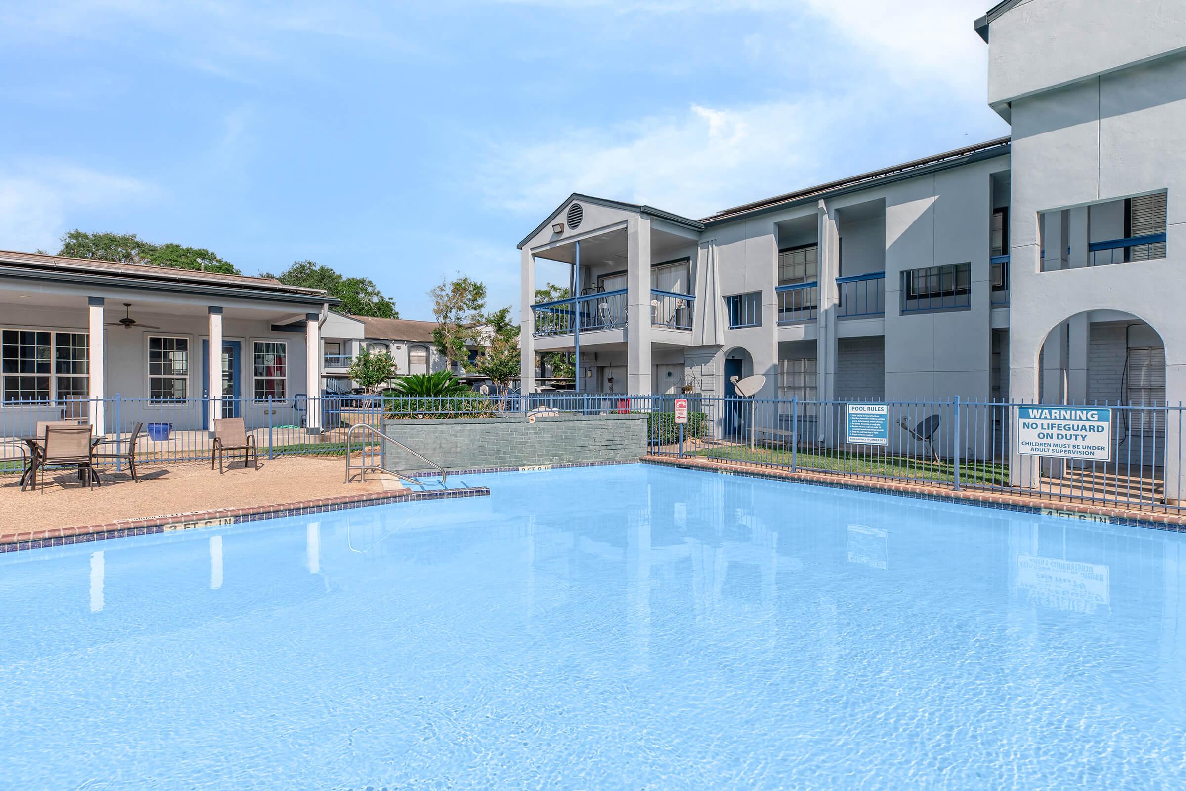 A clear, blue swimming pool surrounded by a patio with lounge chairs and tables. In the background, a multi-story building with balconies and a well-maintained courtyard. Signs are visible near the pool area, indicating safety rules. The scene is bright and inviting, set against a clear sky.