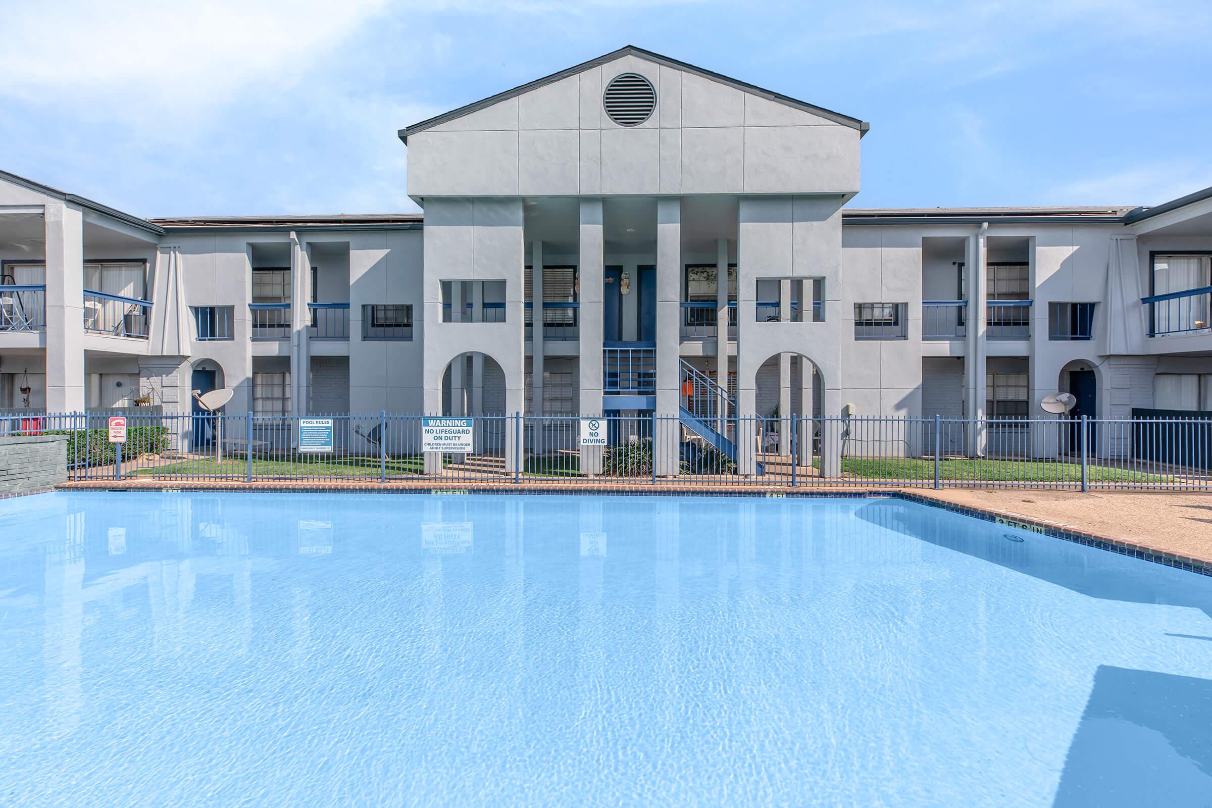 A clear blue swimming pool in the foreground, reflecting a light blue sky. In the background, a modern, multi-story building with a white exterior, featuring several balconies and an entrance with a blue door. The area is surrounded by a security fence, with signs visible nearby.