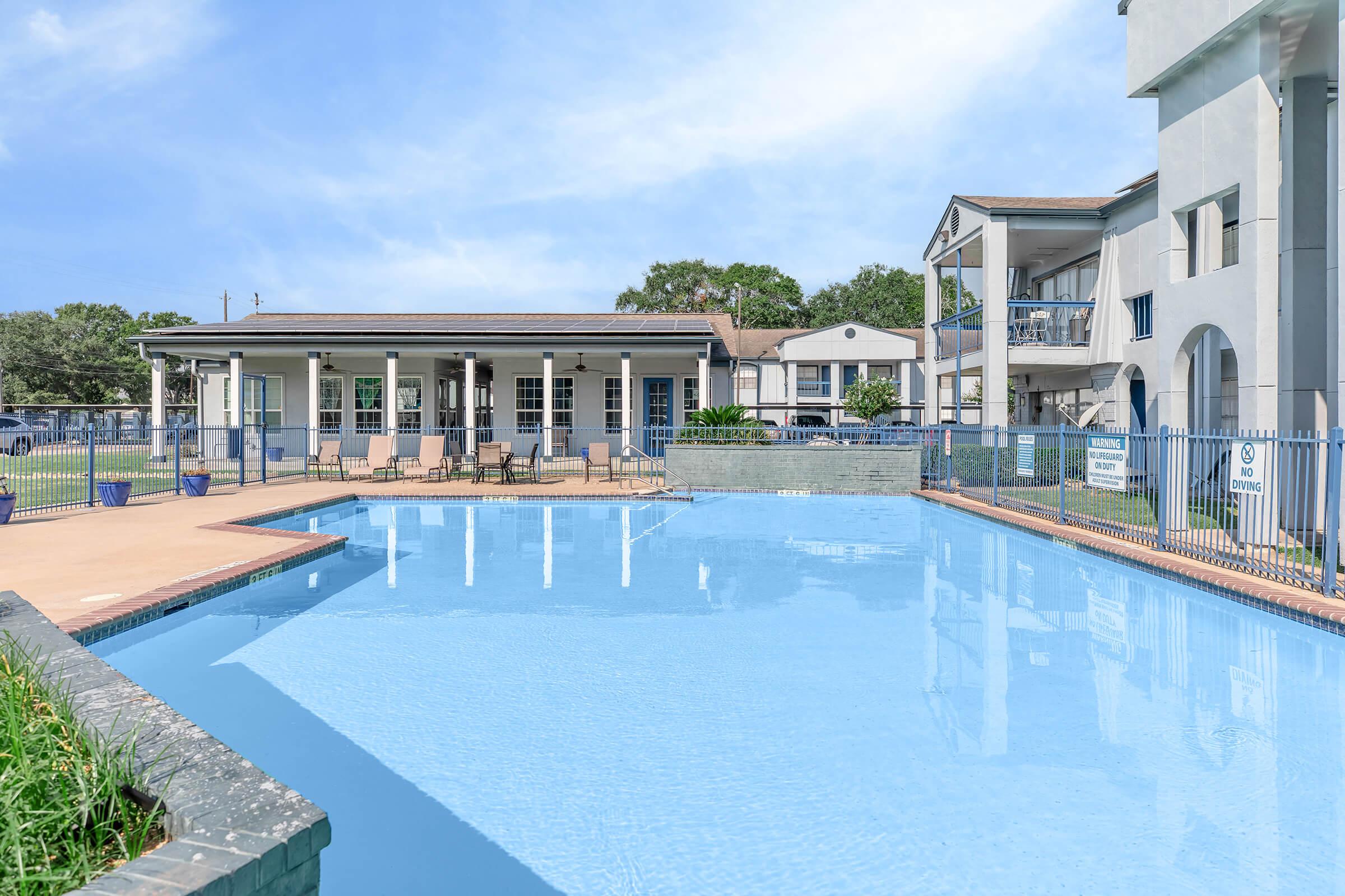 A clear blue swimming pool surrounded by a concrete deck and lounge chairs. In the background, there is a single-story building with multiple windows and a green lawn. The sky is bright and partly cloudy, adding to the inviting atmosphere of the pool area.