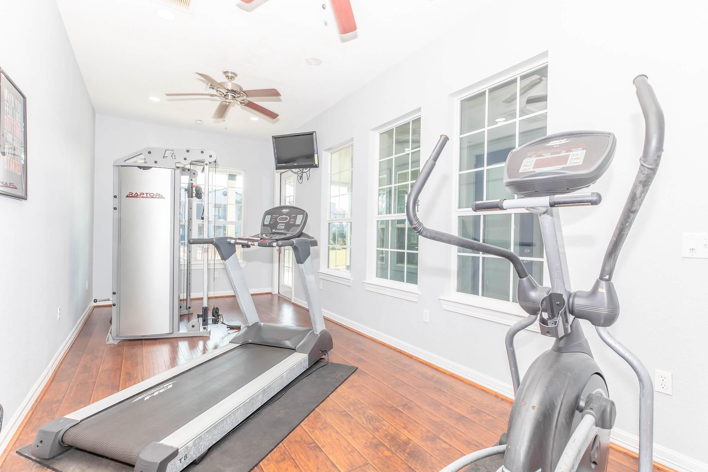 A bright fitness room featuring a treadmill, elliptical machine, and a multi-gym setup. Large windows allow natural light, and a ceiling fan provides ventilation. A television is mounted on the wall for entertainment during workouts. The floor is wooden, creating a warm atmosphere.
