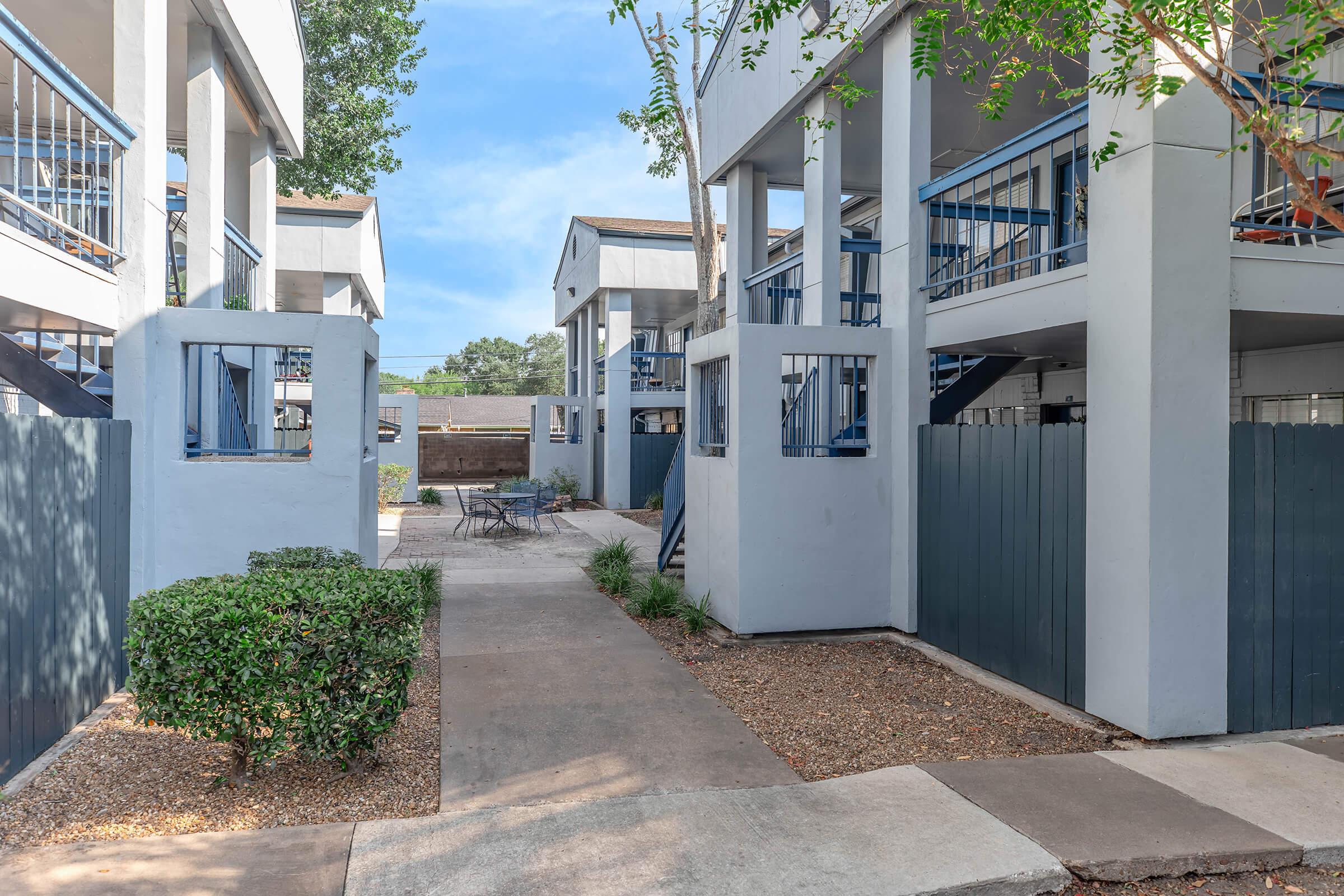Exterior view of a residential complex featuring two-story buildings with balconies. Pathways and green landscaping provide access to the units. A small seating area with outdoor furniture is visible, surrounded by trees and bushes, creating a welcoming and spacious atmosphere.