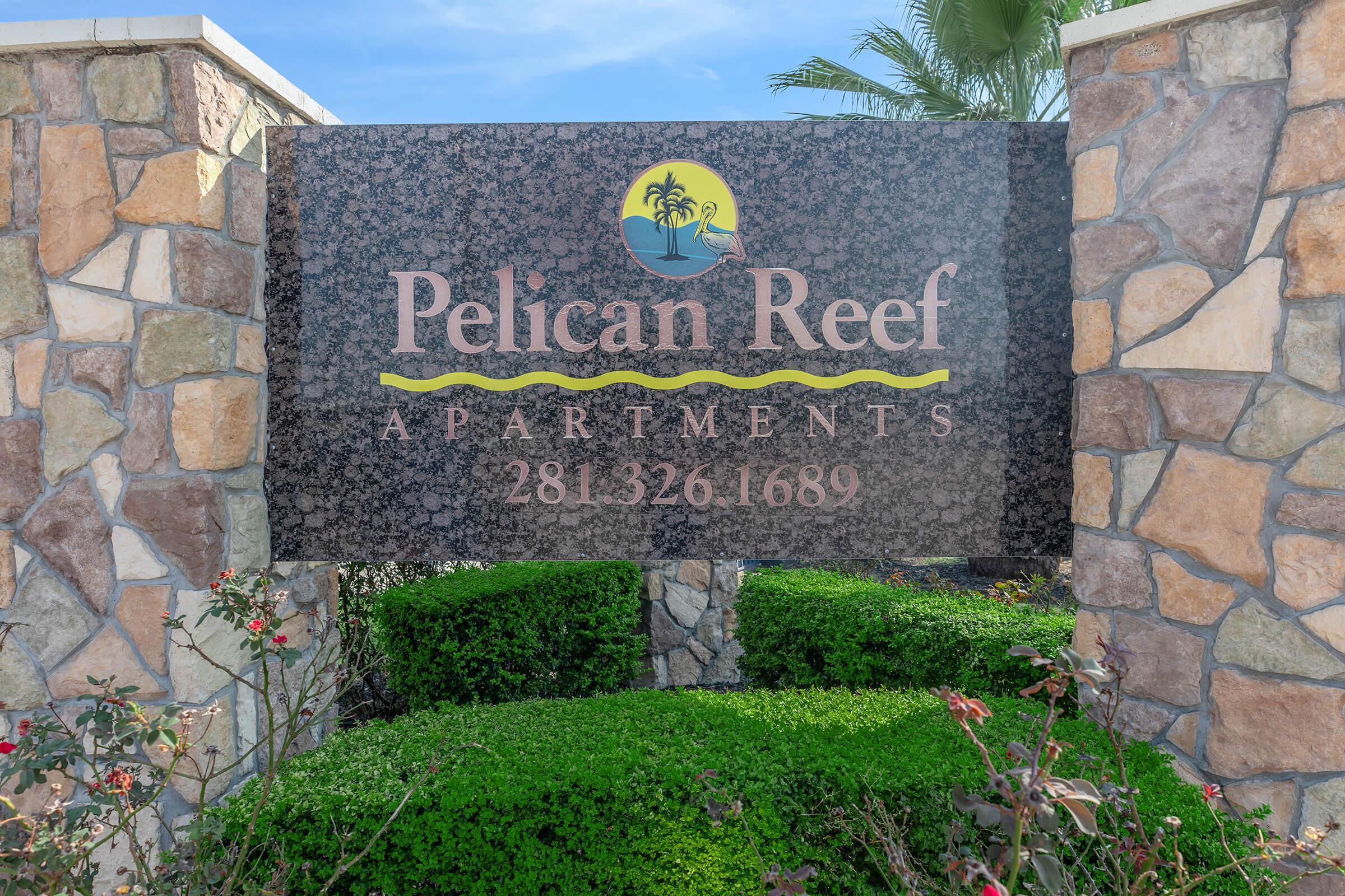 Sign for Pelican Reef Apartments featuring the name prominently displayed in stylized lettering, along with a phone number. The sign is set against a stone background with greenery and palm trees visible, suggesting a tropical environment.