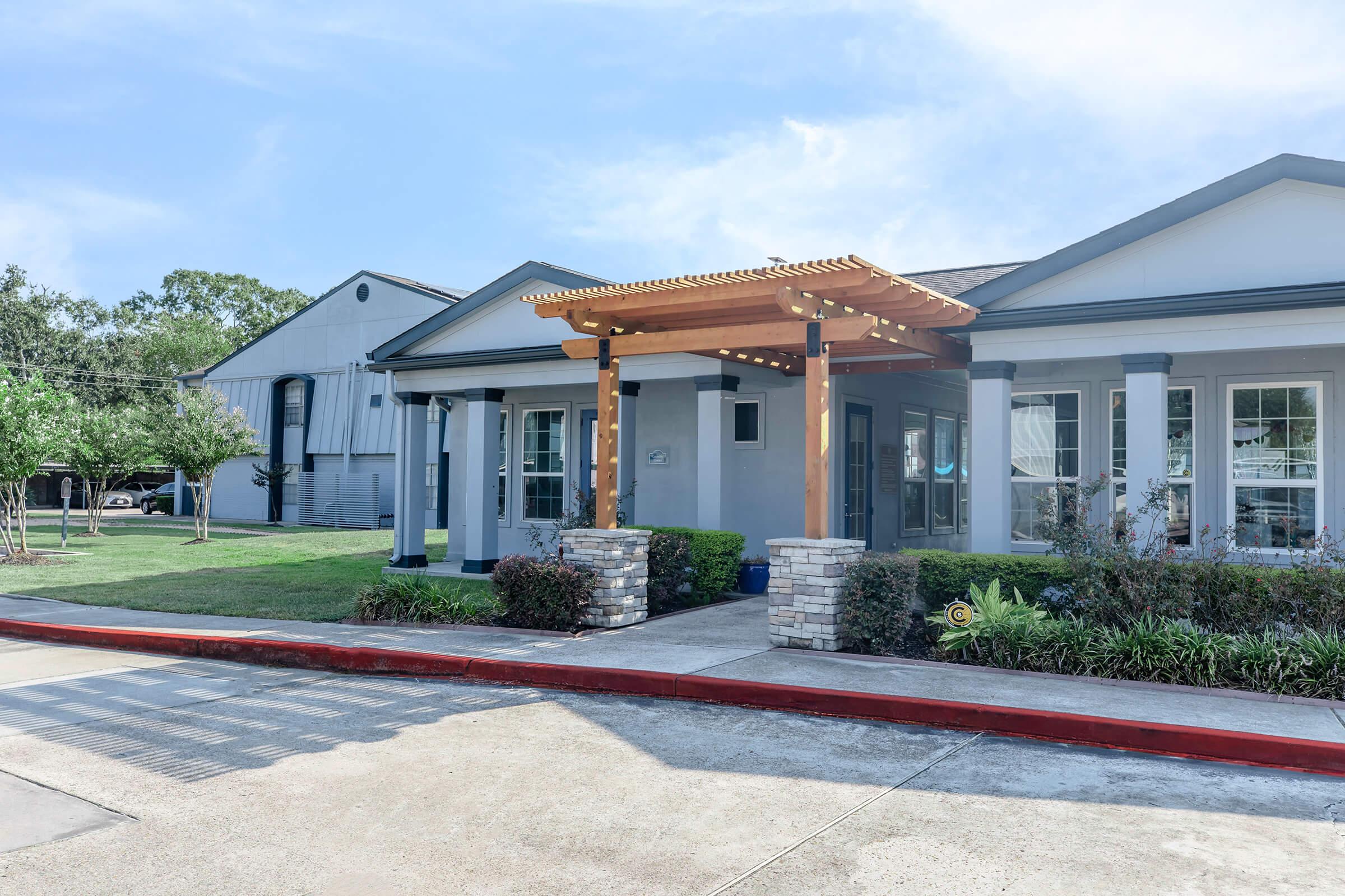 A modern one-story building with a stone entrance and wooden pergola. The facade features large windows and light-colored siding, complemented by well-maintained landscaping. In the background, there is another structure with a metal roof, surrounded by green grass and trees.