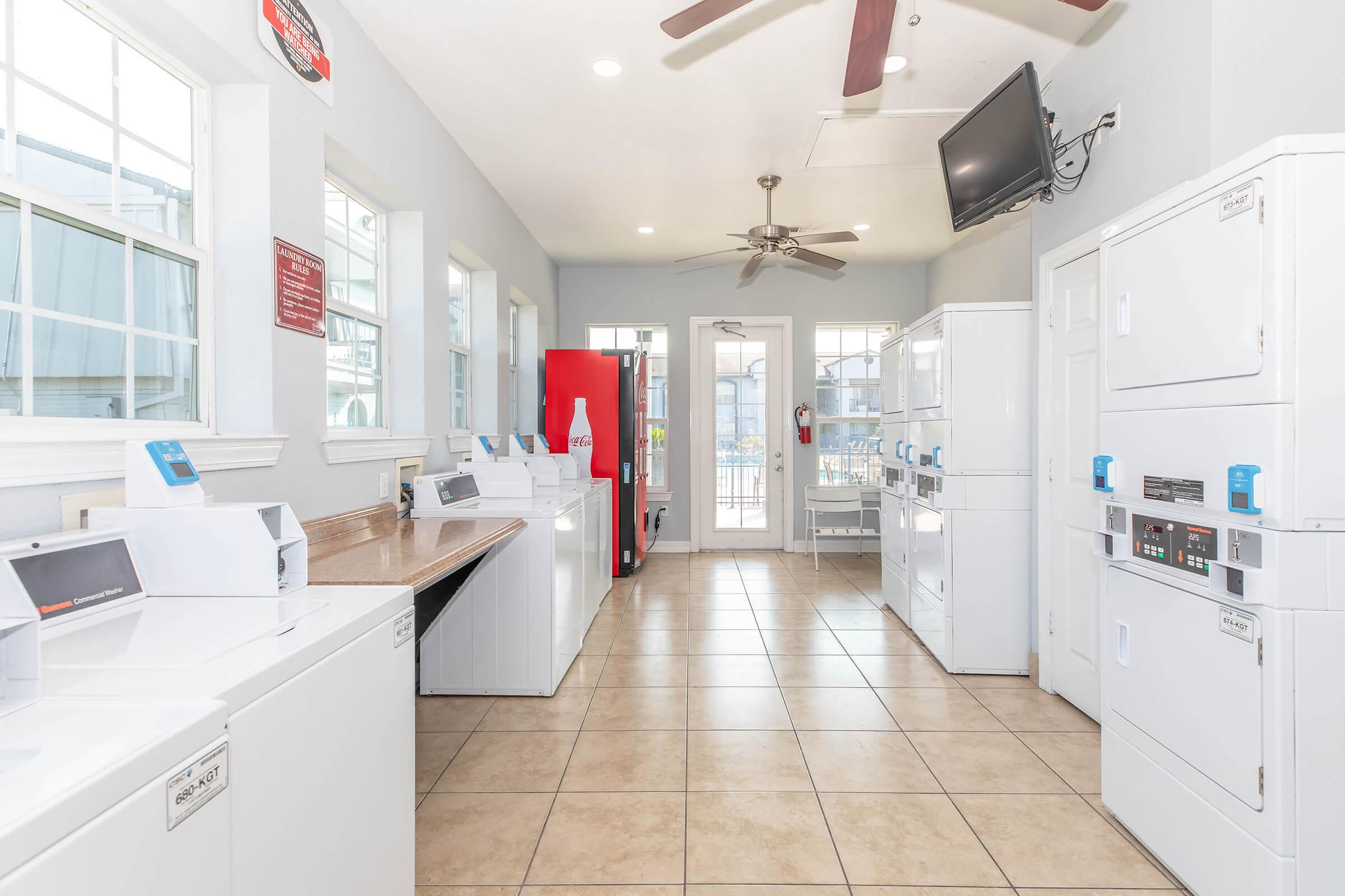 A clean and well-lit laundry room featuring multiple white washing machines and dryers against light-colored walls. Large windows allow natural light, and there is a red vending machine, a ceiling fan, and a TV mounted on the wall, providing a functional and comfortable space for laundry activities.