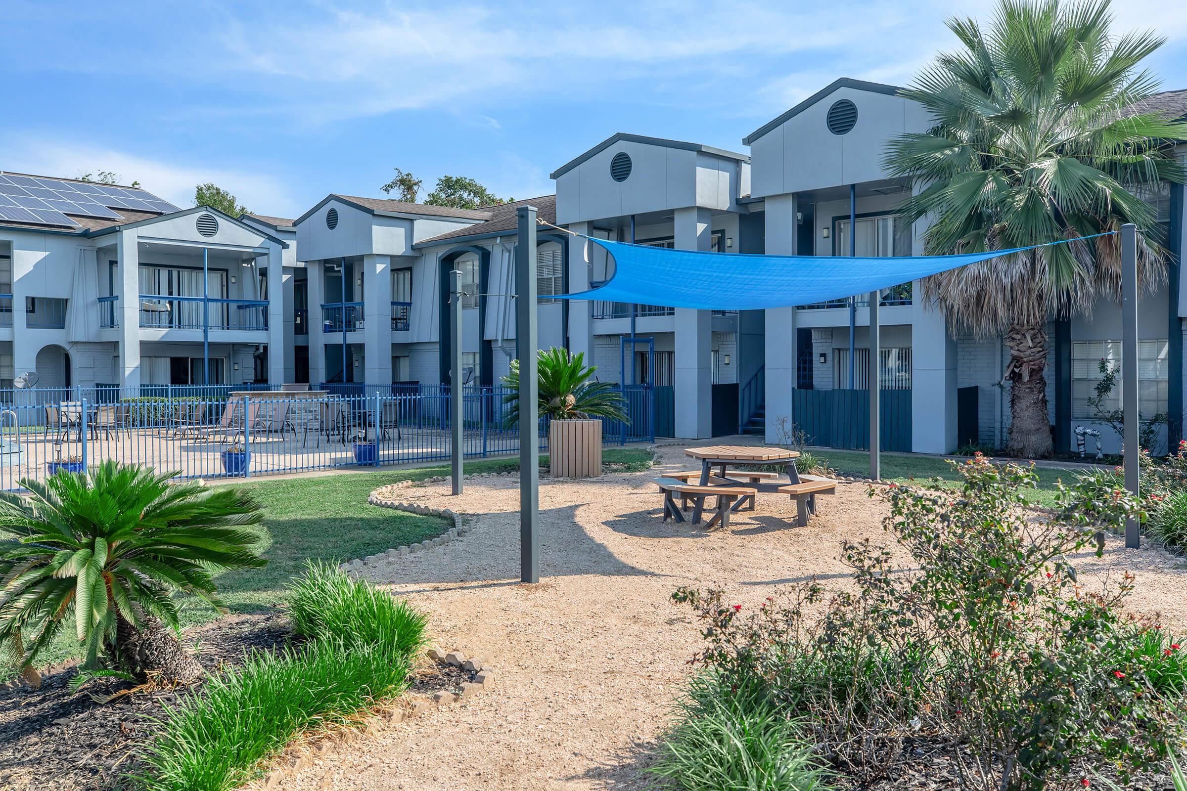 A landscaped area with a picnic table and blue shade sail in front of modern apartment buildings. The scene features palm trees, well-maintained grass, and playground equipment in the background, creating a welcoming outdoor space.