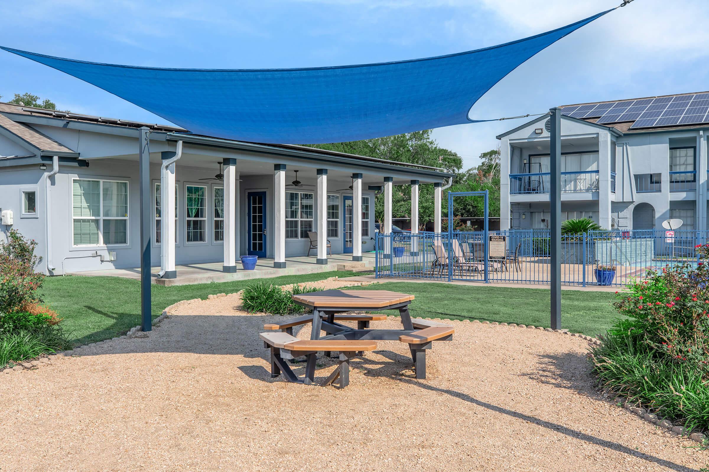 A sunny outdoor area featuring a picnic table under a blue shade sail, surrounded by landscaped gravel and greenery. In the background, there is a residential building with balconies and access to a swimming pool, with solar panels on the roof.