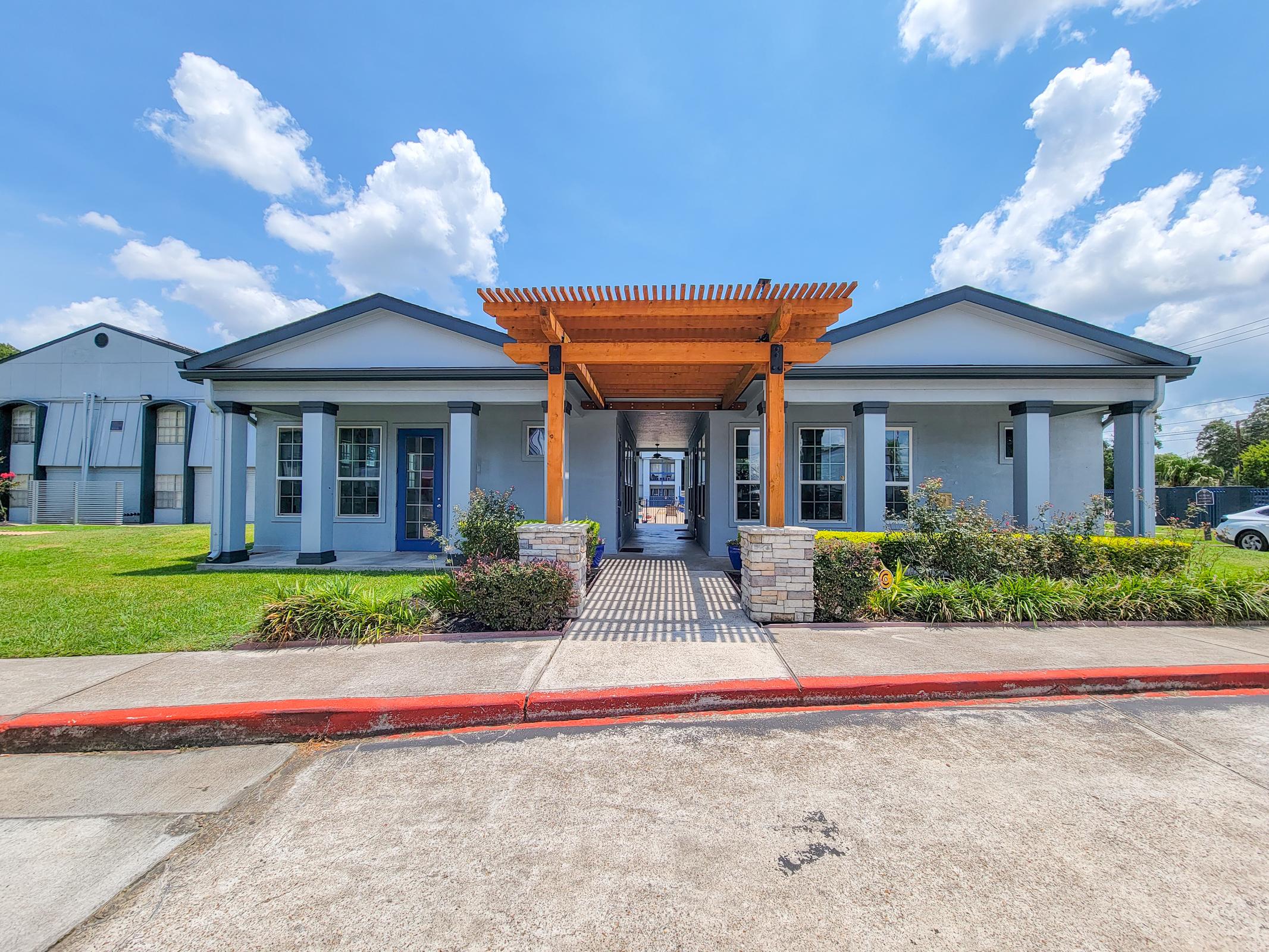 A modern one-story building with a front porch featuring wooden beams, surrounded by green grass and landscaping. The sky is bright blue with fluffy white clouds, and the pathway leading to the entrance is shaded by the porch structure.