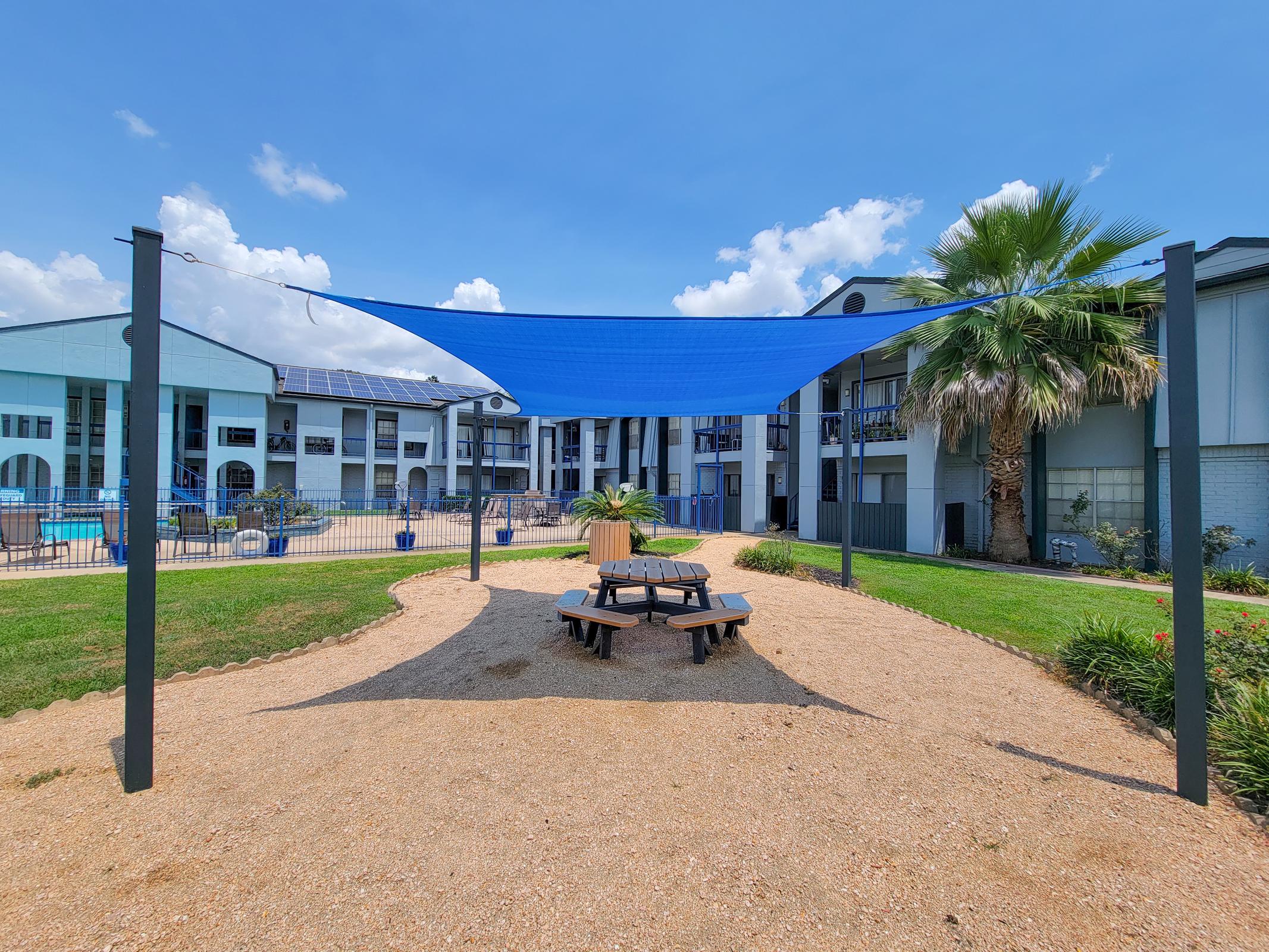 A shaded seating area with a blue canopy over a picnic table in a gravelly park setting. Surrounding the area are green lawns and palm trees, with a backdrop of a multistory building featuring balconies and solar panels under a bright blue sky.