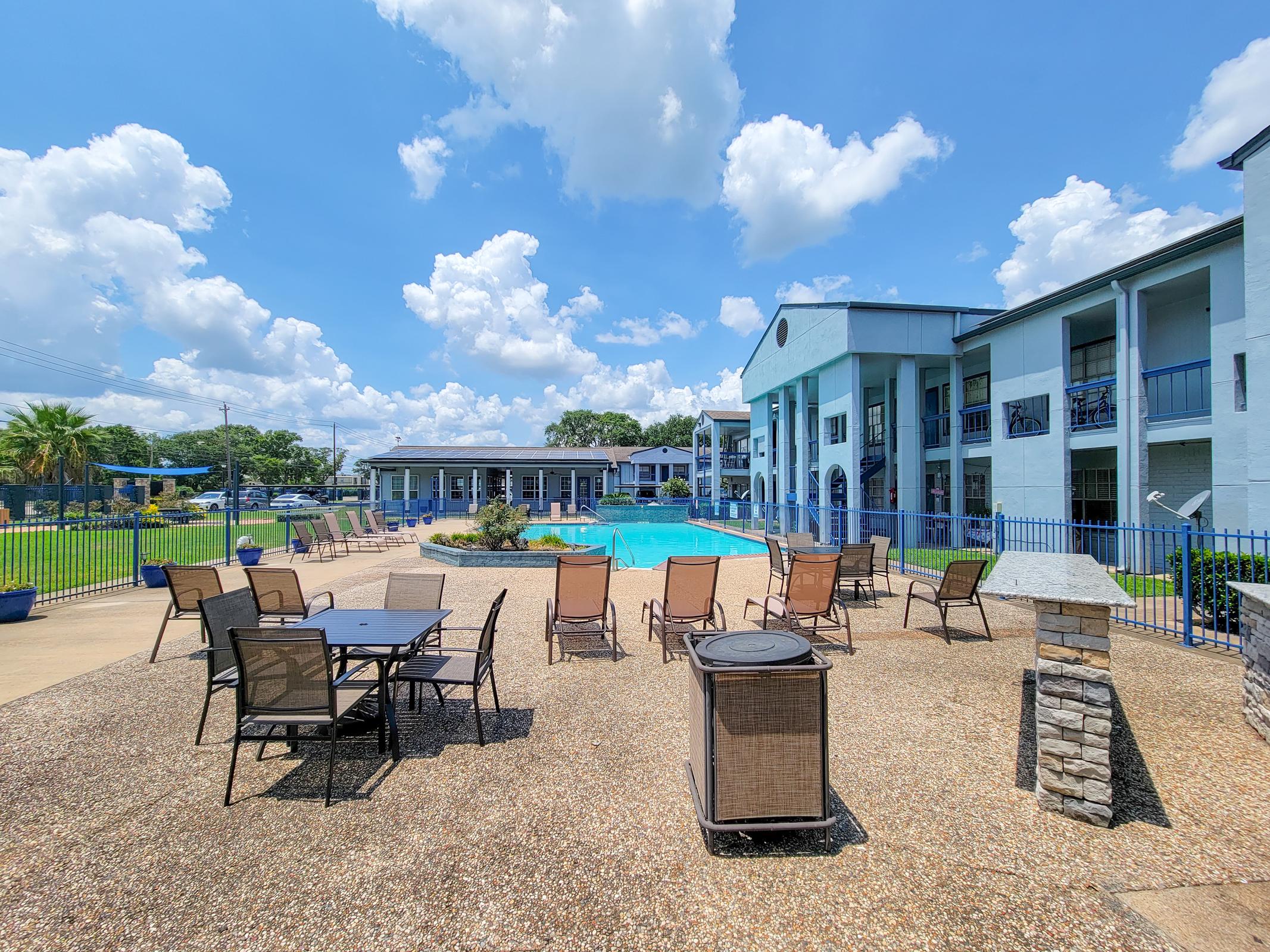 A sunny outdoor area featuring a swimming pool surrounded by lounge chairs and tables. The setting includes a well-maintained lawn, palm trees, and a blue sky with fluffy clouds. Nearby buildings are partially visible, adding to the inviting atmosphere of the poolside space.