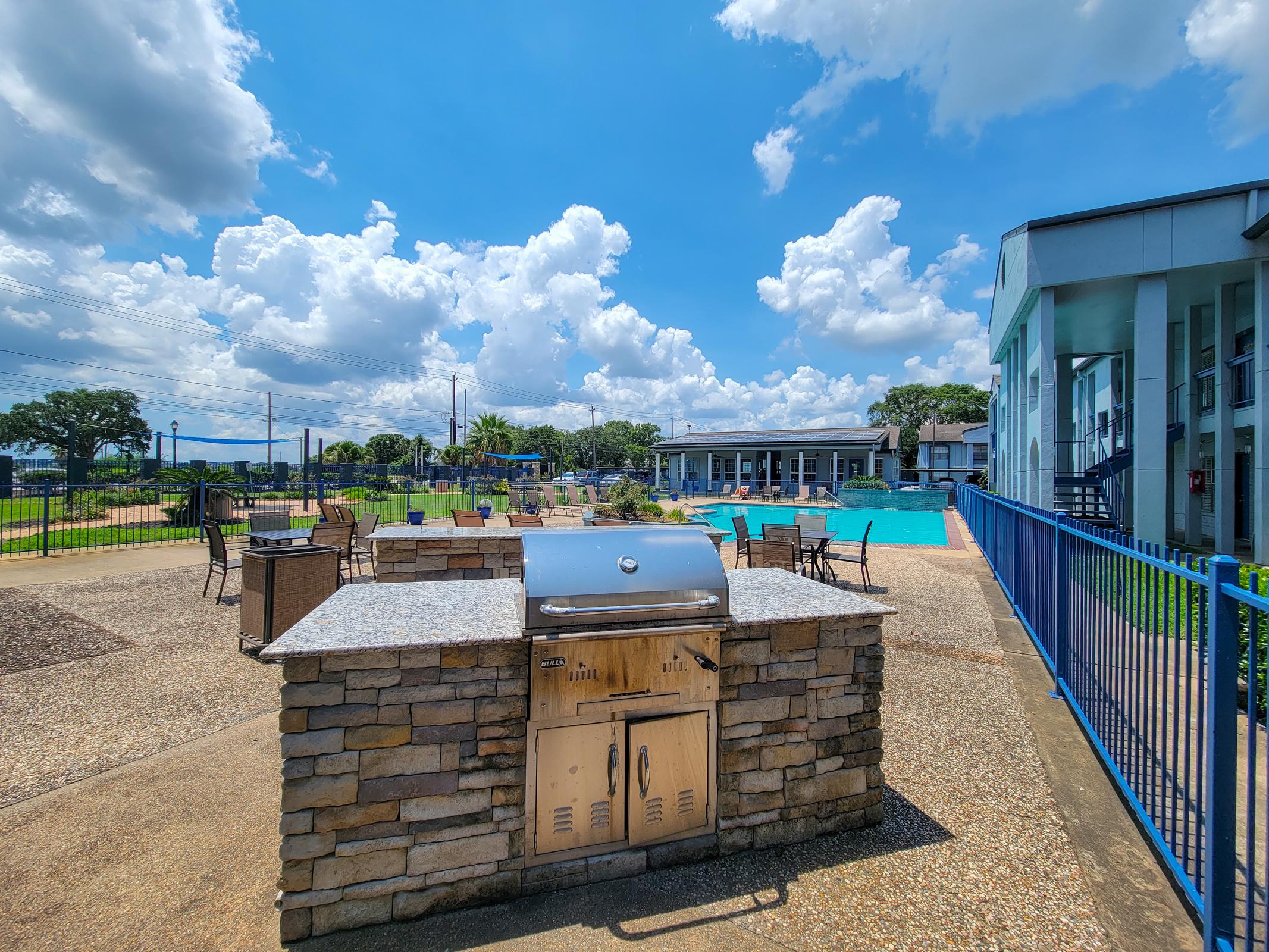 Outdoor barbecue area with a stone grill, surrounded by tables and chairs, adjacent to a swimming pool. The background features a blue sky with fluffy white clouds and a well-maintained green lawn. The scene is bright and inviting, suggesting a leisure or recreational space.