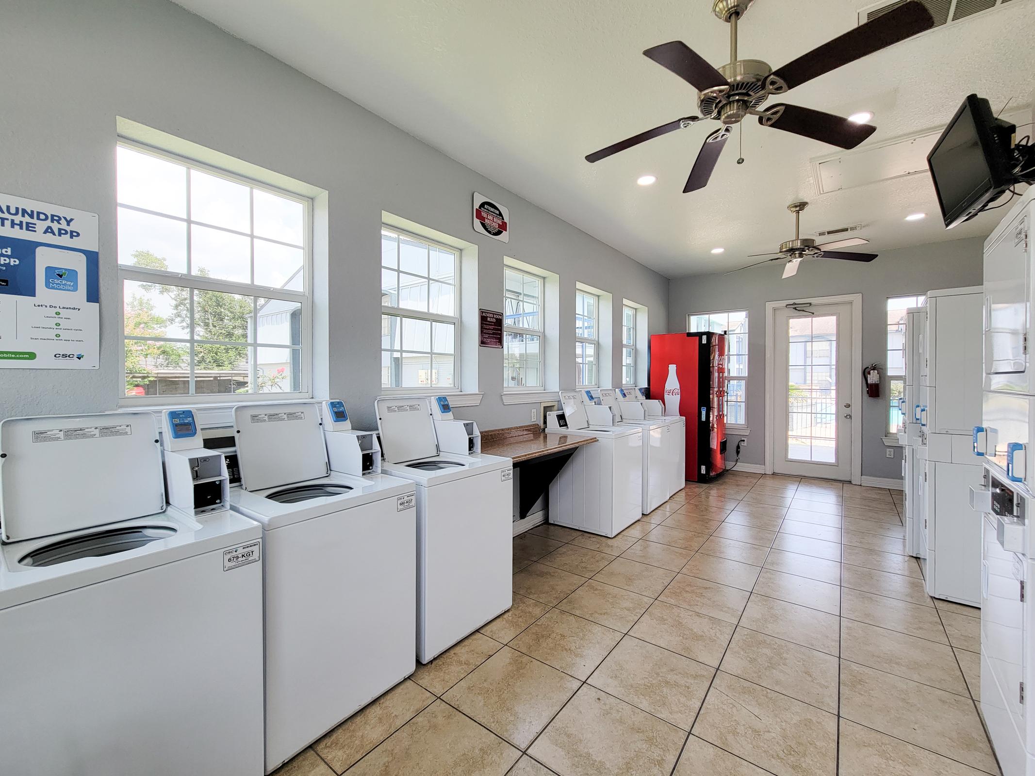 A clean and bright laundry room featuring several white washing machines and dryers lined up against the wall. Large windows let in natural light, and there's a drinking vending machine in the corner. The space is well-maintained with tiled flooring and ceiling fans for ventilation.