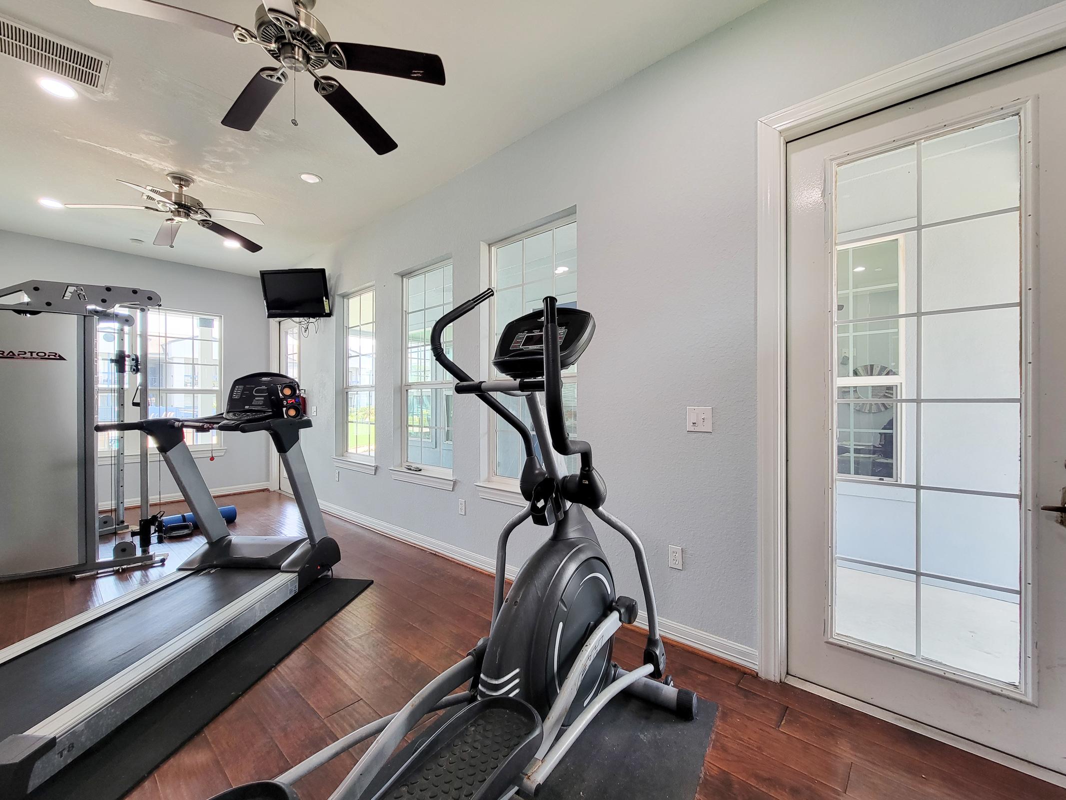 A bright home gym featuring a treadmill and an elliptical machine, with large windows allowing natural light. The gym has a ceiling fan and a door leading to another room, creating an open and airy atmosphere. The wooden floor adds warmth to the modern space.