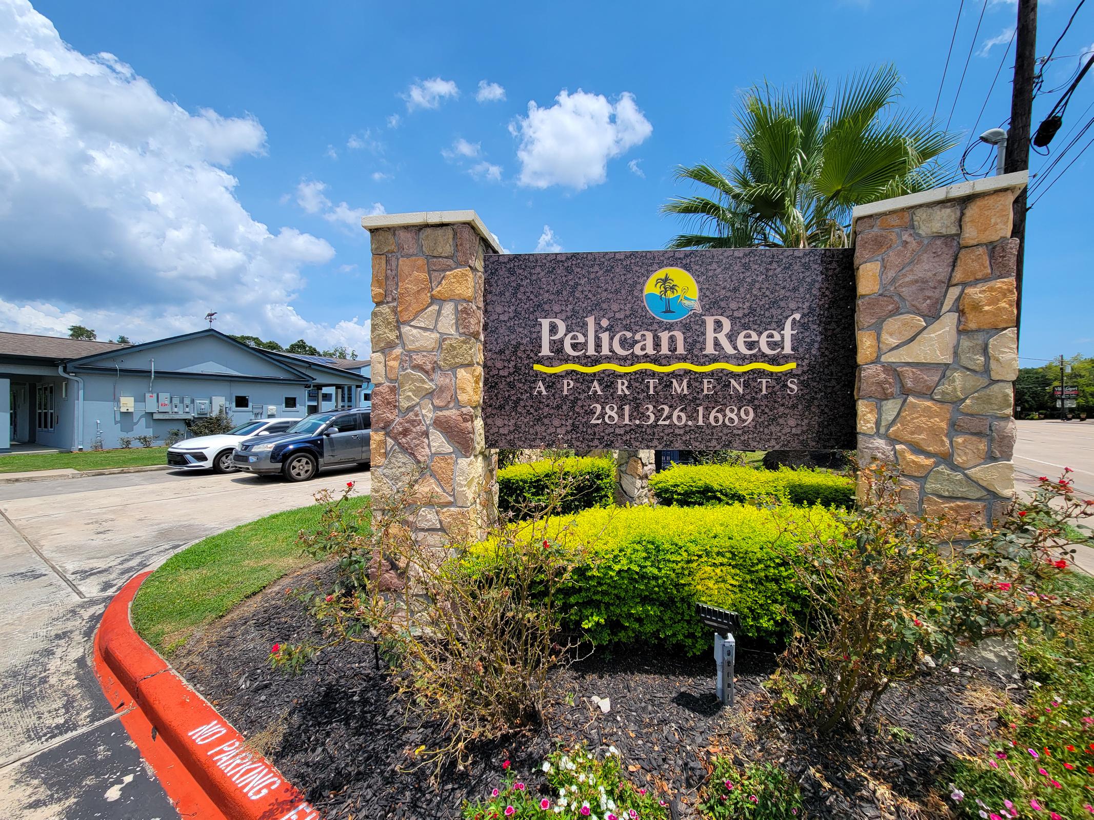 Sign for Pelican Reef Apartments with a decorative stone base, featuring a palm tree logo. The area is landscaped with flowers and shrubs, and a parking area can be seen. Light blue sky with some clouds in the background.