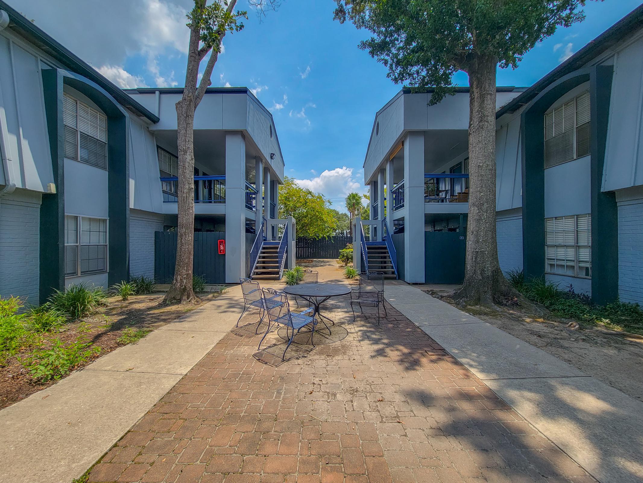 A view of a landscaped courtyard between two multi-story apartment buildings. There are two trees and a paved pathway leading to stairs on either side. A round table with chairs is situated in the center of the courtyard, surrounded by grass and shrubs, under a clear blue sky with a few clouds.