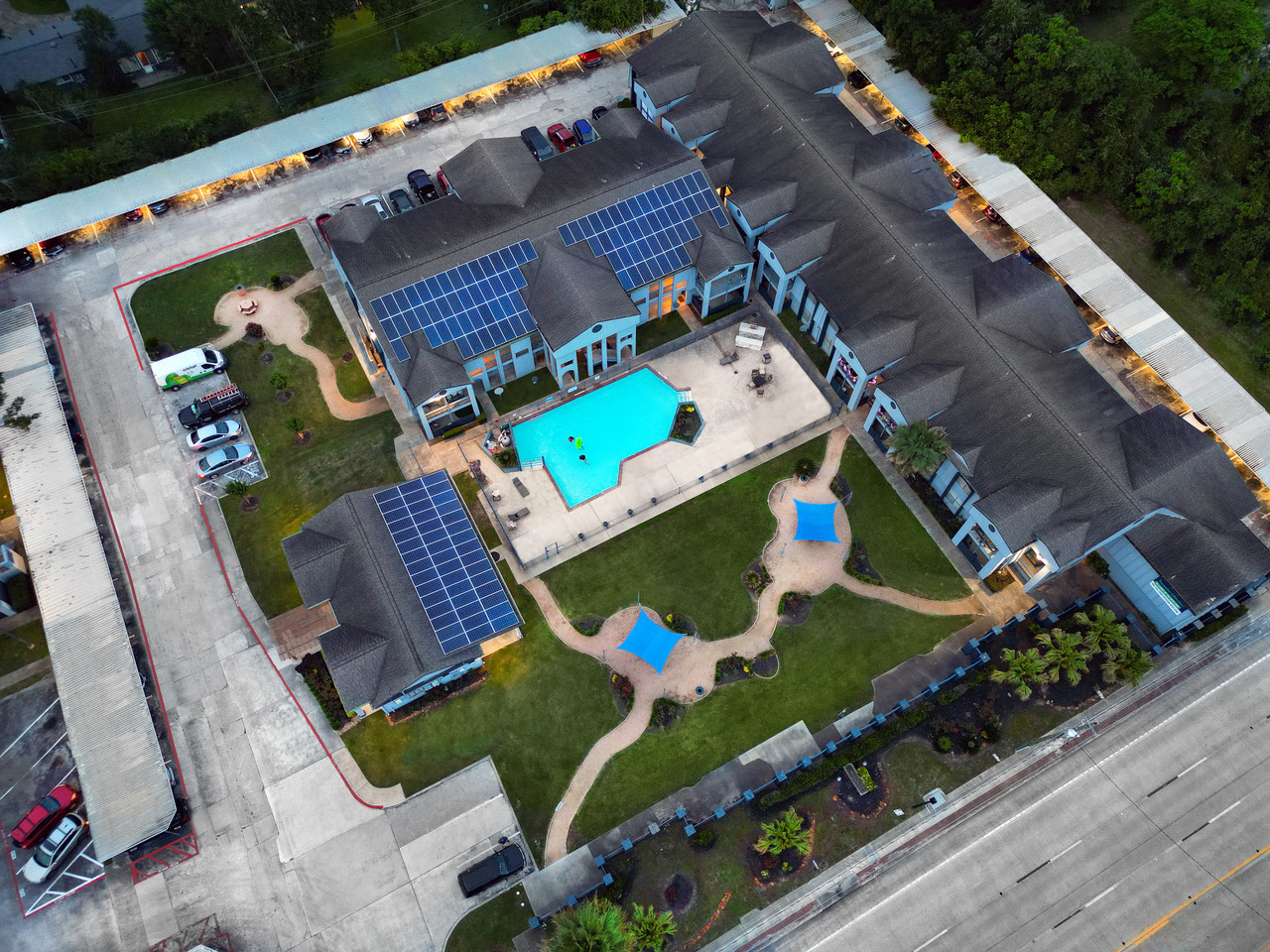 Aerial view of a residential building complex featuring a central pool surrounded by landscaped grass, lounge areas, and palm trees. Solar panels are visible on the rooftops, and parking spaces line the perimeter. The setting is serene and inviting, ideal for relaxation.