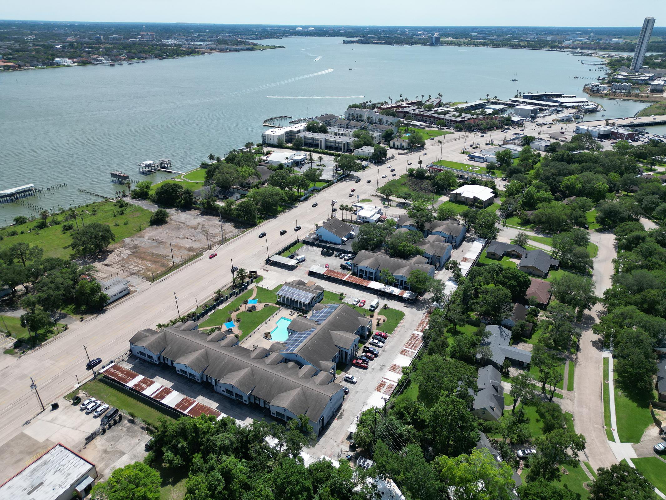 Aerial view of a waterfront area featuring a river and marina, lined with buildings and roads. The landscape includes a mix of residential and commercial structures, green spaces, and a hotel with a pool, with vehicles on the nearby road and trees surrounding the properties.