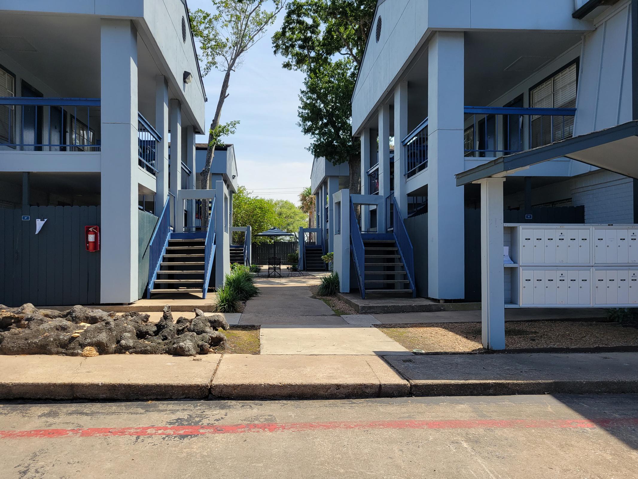 A view of a residential area featuring two buildings with blue accents. Stairs lead up to individual units on either side of a paved walkway. There are mailboxes mounted on a wall, and greenery, including small trees and shrubs, enhances the scenery. The sky is clear, suggesting a sunny day.