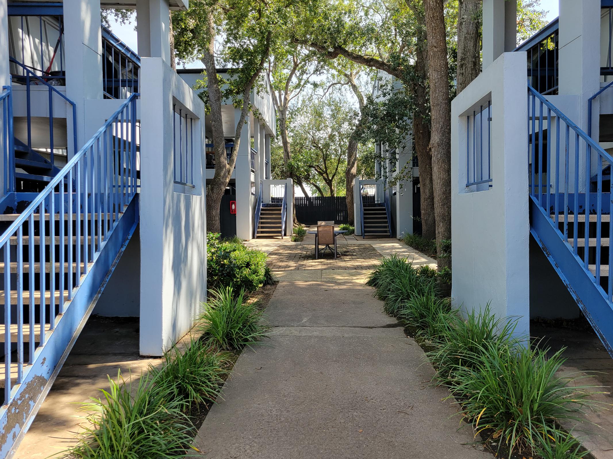 A walkway between two buildings featuring blue railings and stairs leading to upper levels. Lush green grass and plants line the walkway, creating a serene atmosphere. A chair is positioned in the middle of the pathway, inviting relaxation. Trees provide shade along the sides.