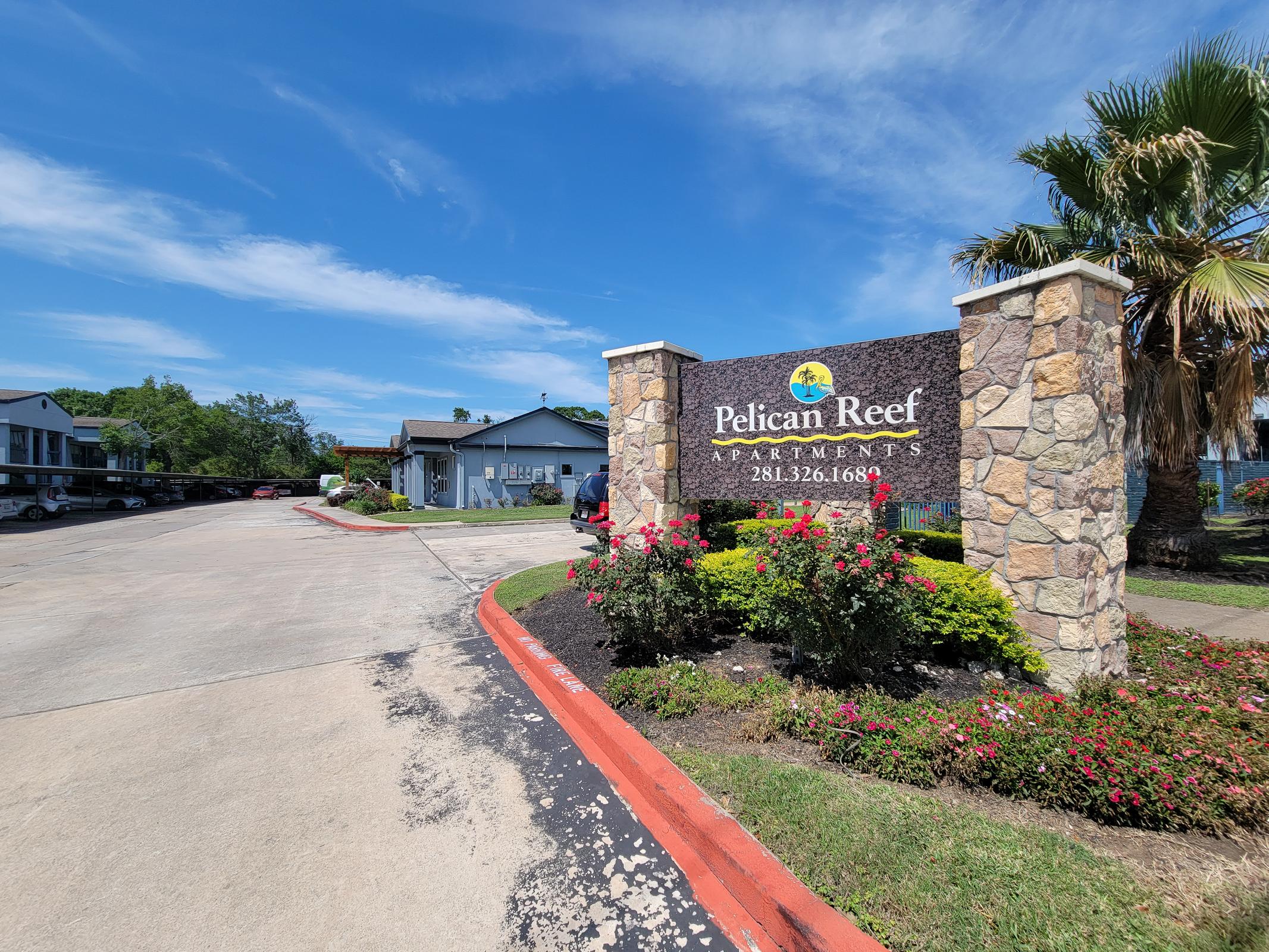 Sign for Pelican Reef Apartments, featuring a stone monument with the name and contact number. The entrance is surrounded by well-maintained landscaping, including colorful flowers and palm trees, under a clear blue sky.