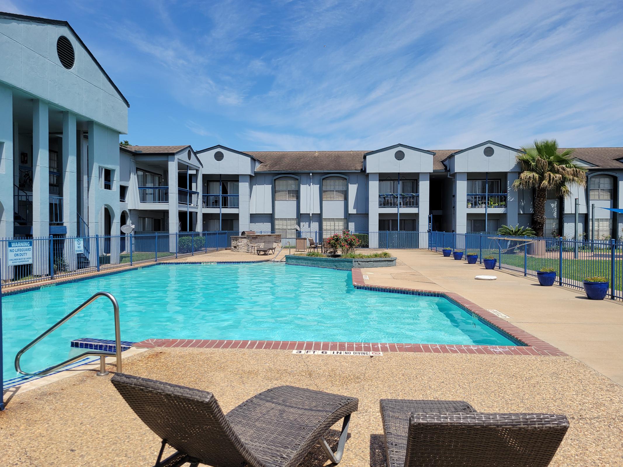 A clear blue swimming pool surrounded by a paved area with two lounge chairs in the foreground. In the background, a multi-story apartment building with blue and gray accents, bordered by green palm trees and landscaping, under a bright blue sky with scattered clouds.