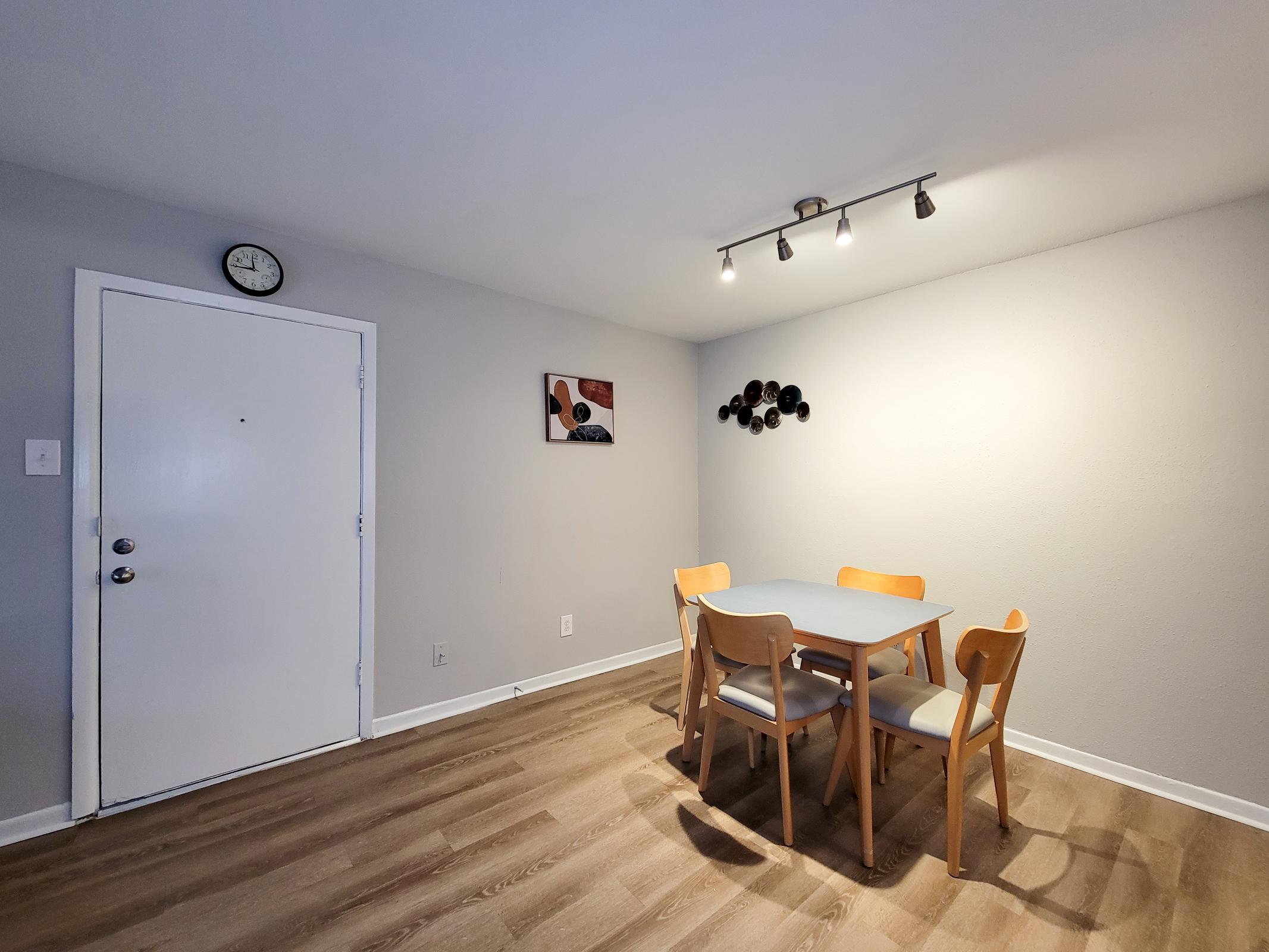 A minimalist dining area featuring a light gray wall, a round table with four wooden chairs, and a wall clock. Sunlight illuminates the space, with modern artwork on the wall and decorative circular wall art. The floor is finished with light wood planks, creating a warm ambiance.
