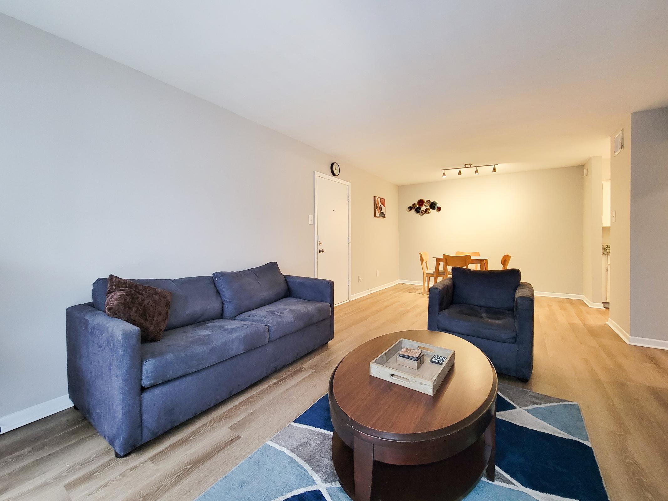 A cozy living room featuring a blue couch and a matching chair, a wooden circular coffee table on a colorful area rug, and a dining area in the background with a table and chairs. The walls are painted light gray, and there is a clock and artwork on display. Natural light brightens the space.